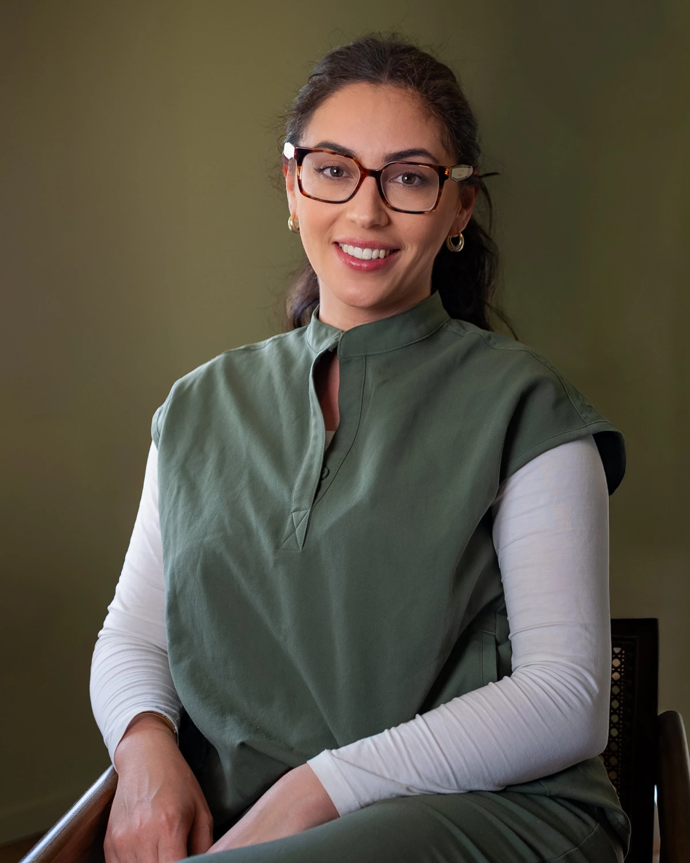 A woman with glasses and earrings smiling, wearing a green vest over a white long-sleeve shirt, sitting on a chair against a green background.
