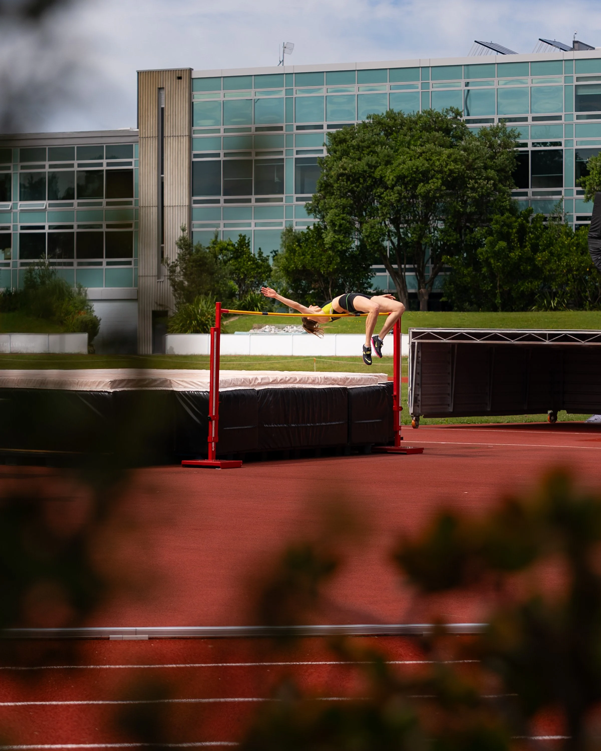 A female athlete in mid-air during a high jump attempt at an outdoor track and field stadium with a modern building and trees in the background.