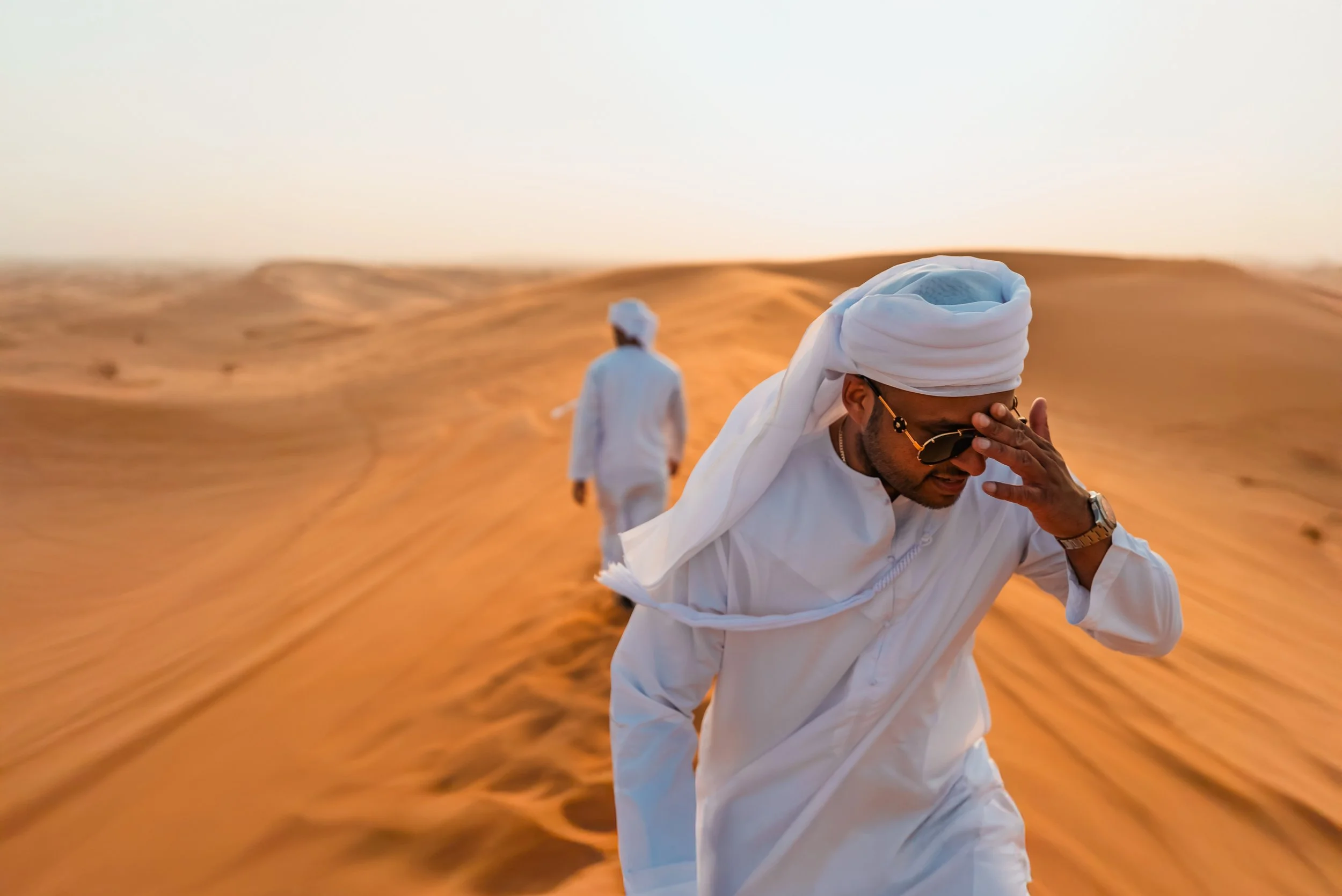 Two men in traditional white clothing walking through a desert landscape at sunset, with one man holding his head and the other walking ahead.