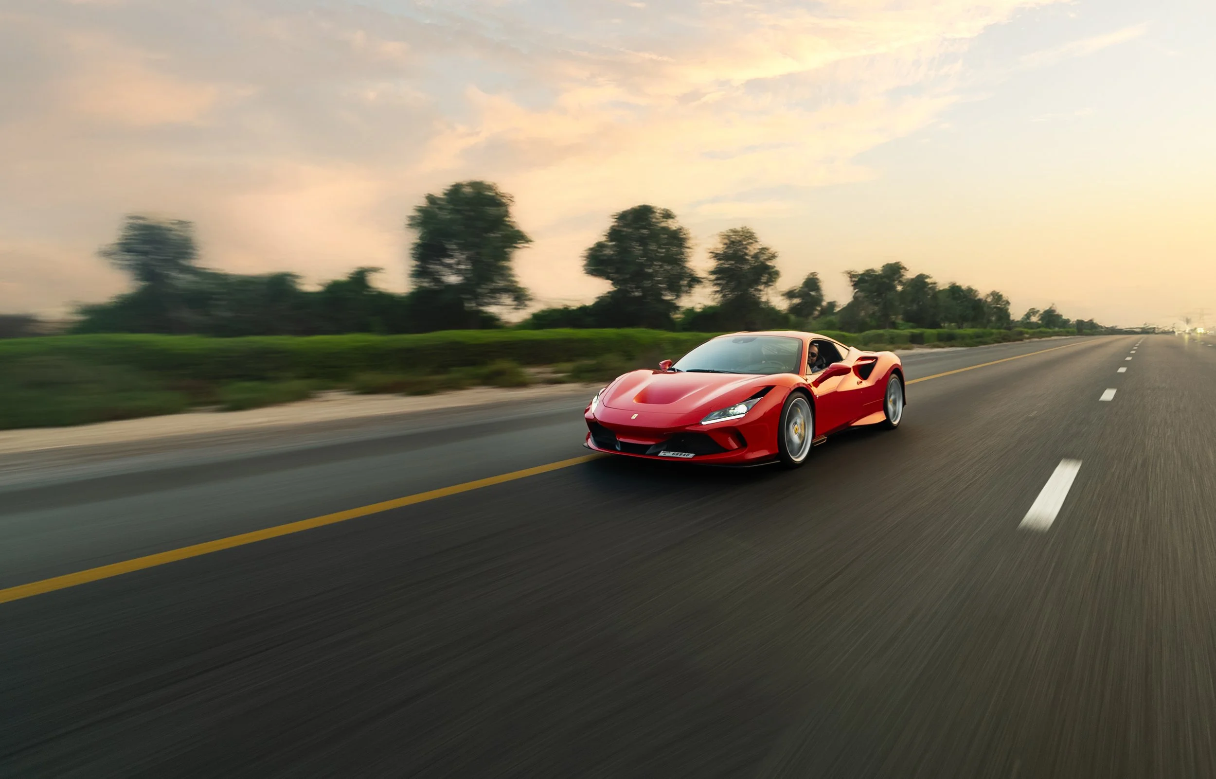 Red sports car driving on an open road with trees in the background during sunset.