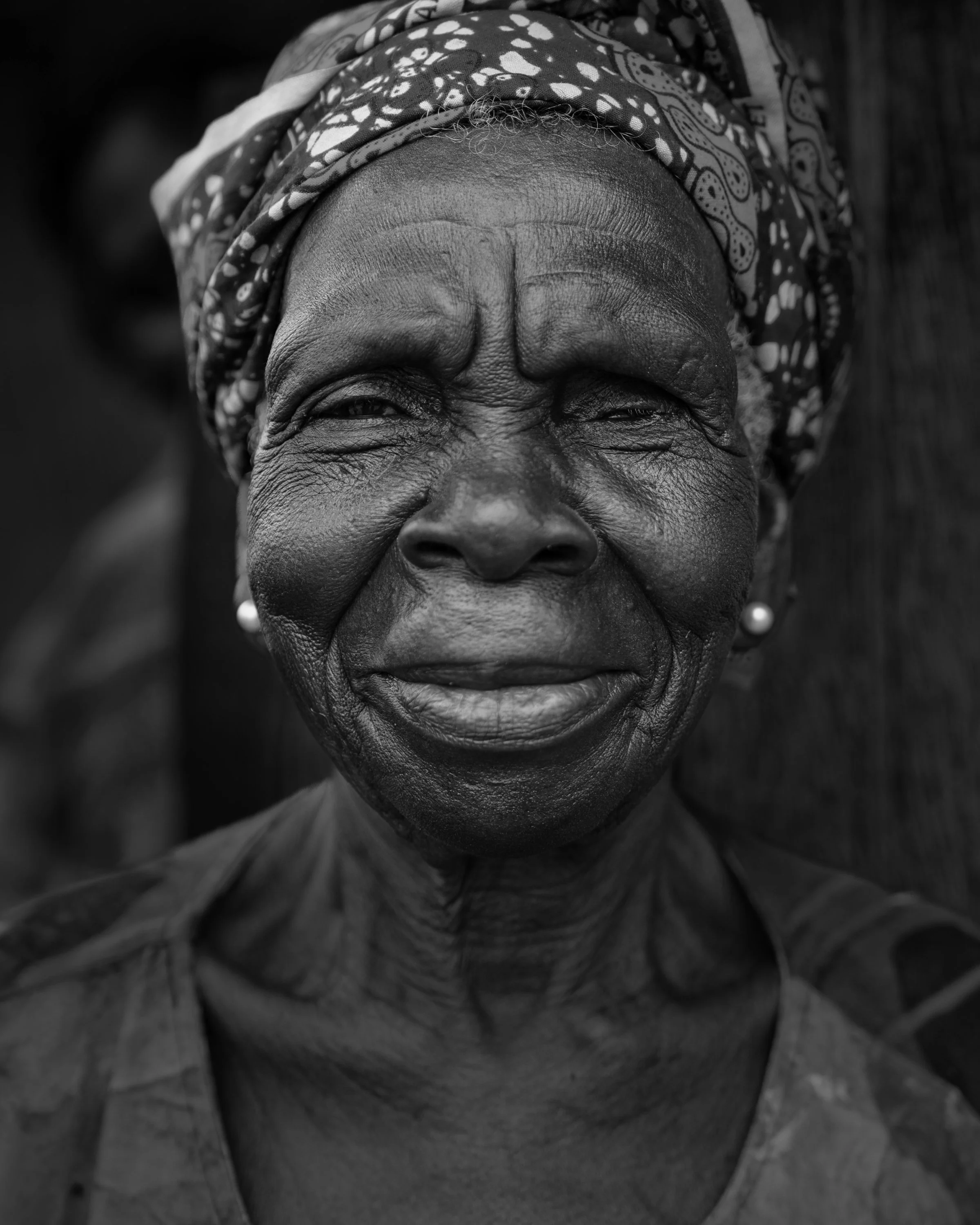 Close-up black and white portrait of an elderly woman with a patterned headscarf and earrings, smiling gently.