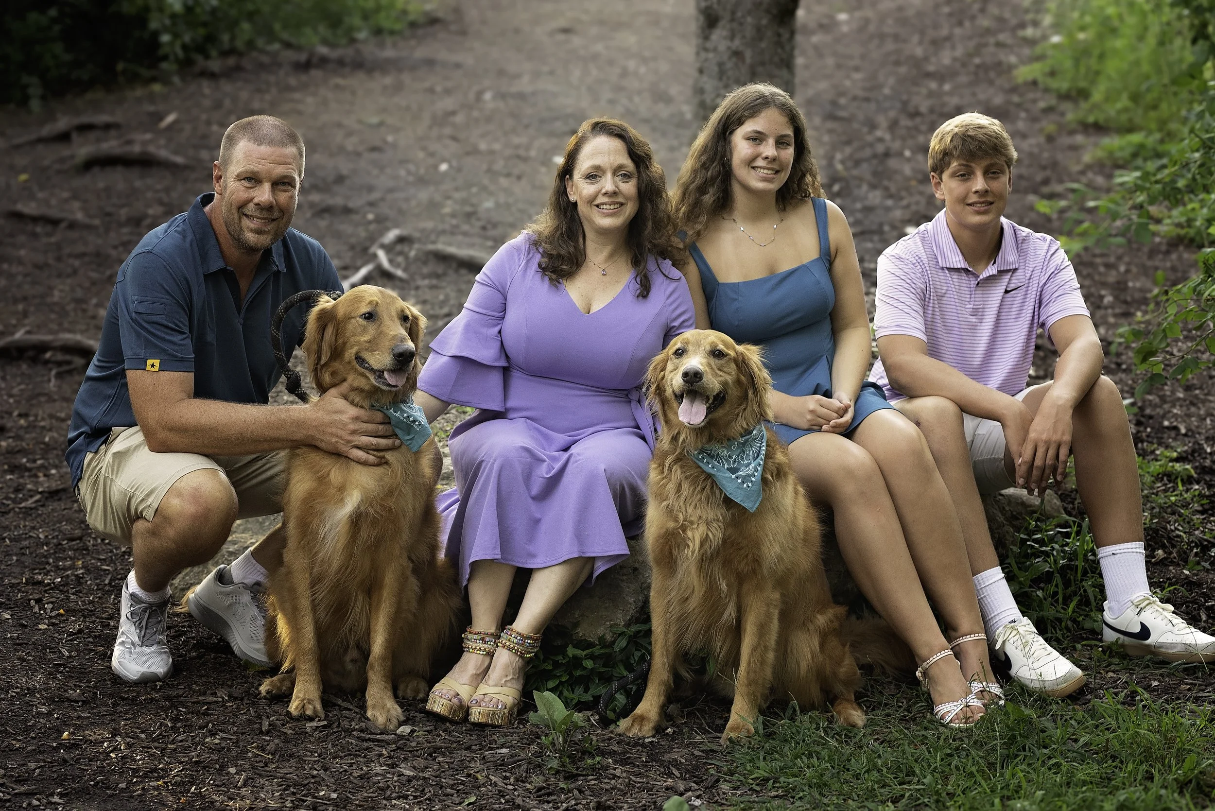 Family of four with two golden retrievers sitting outdoors in a wooded area.