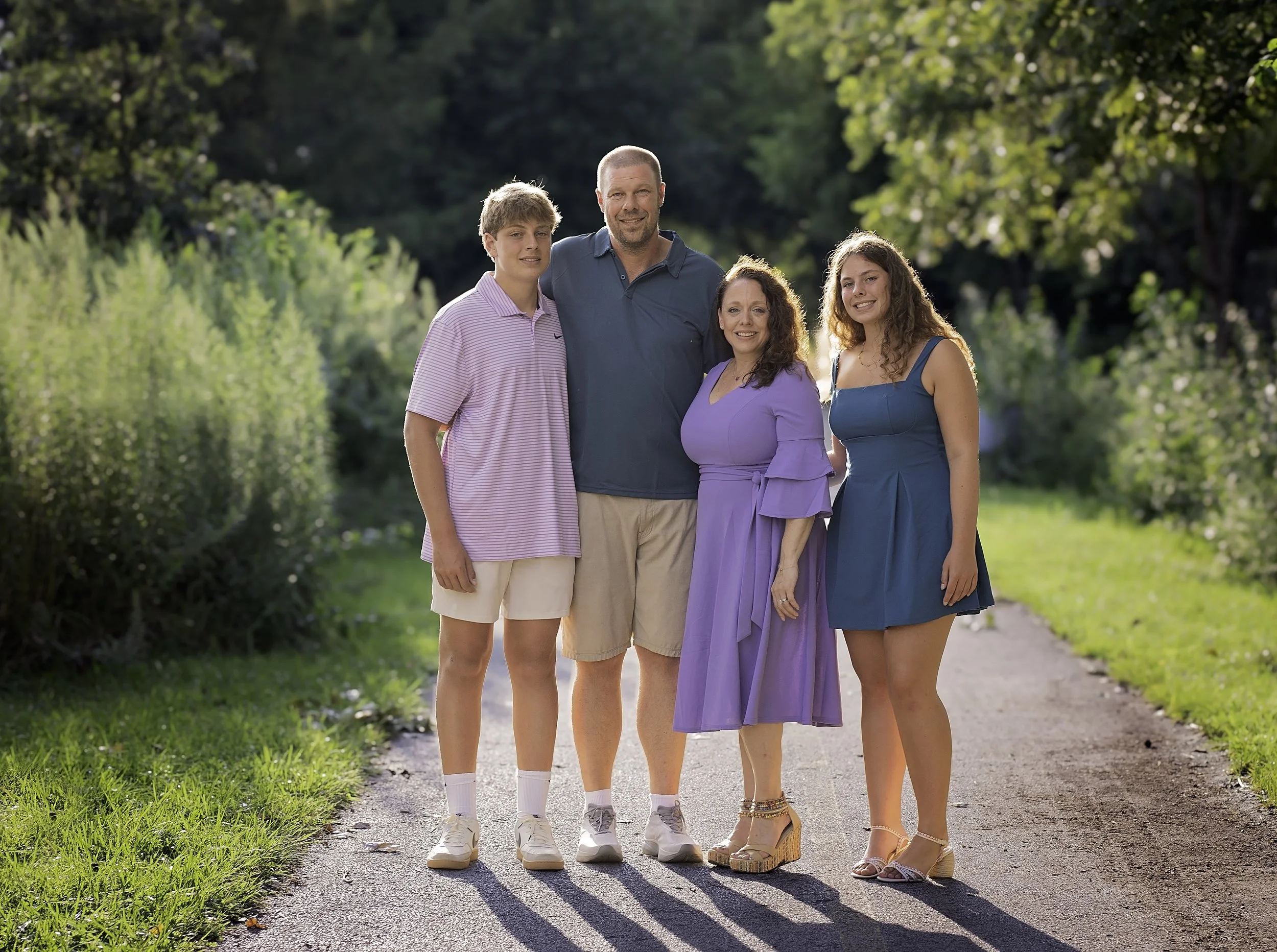 Family of four standing outdoors on a sunny day, smiling, with trees and greenery in the background.