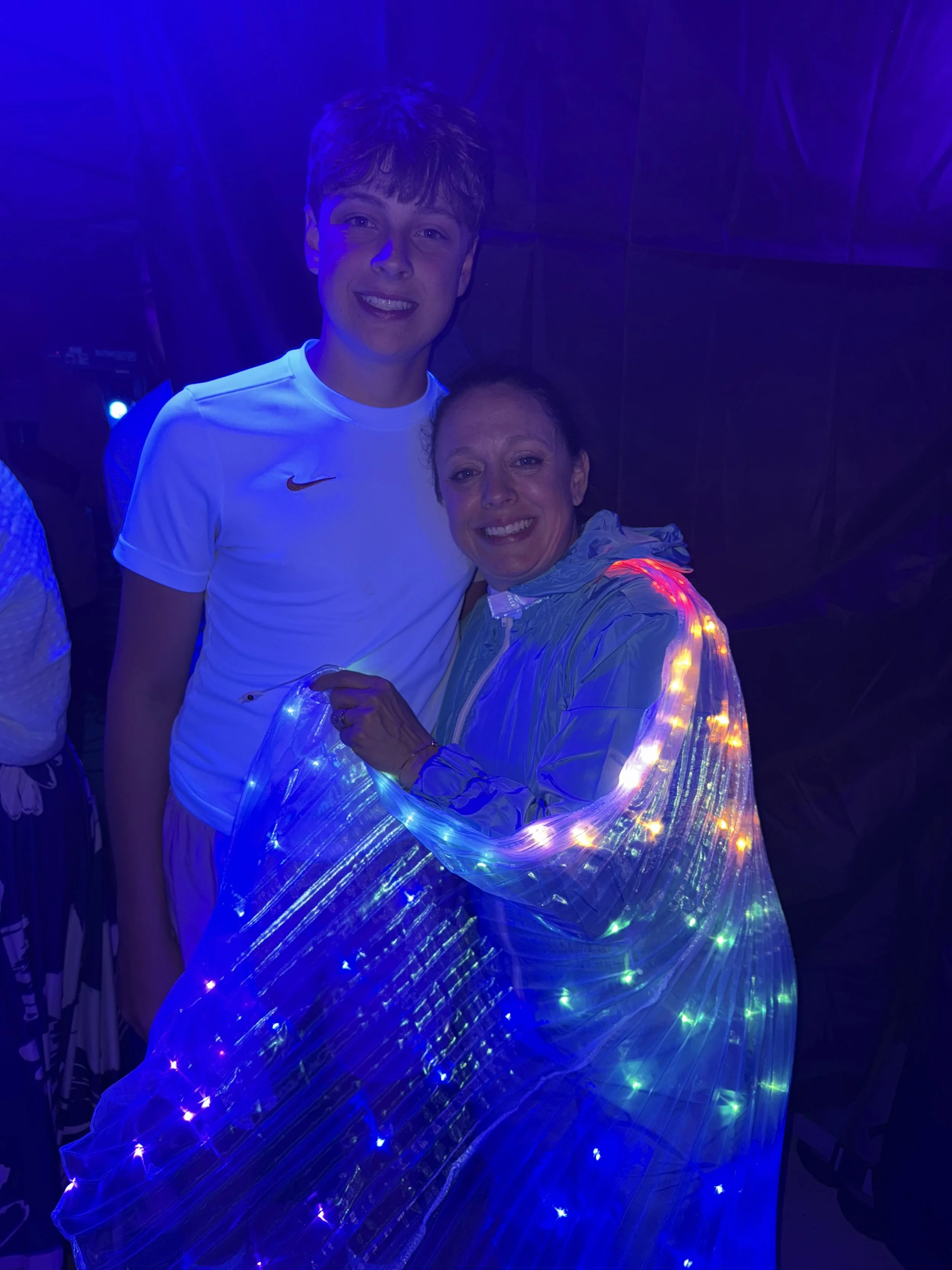A young man and a woman smiling, standing close together at a dark event with blue and multicolored LED lights.