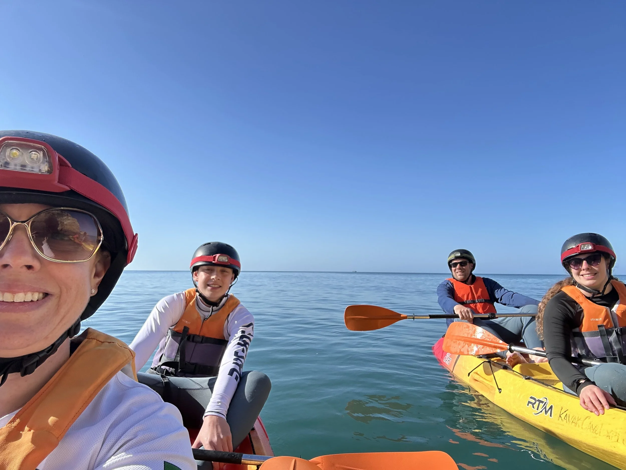 Four people wearing life jackets and helmets kayaking on calm water under a clear blue sky, taking a selfie.