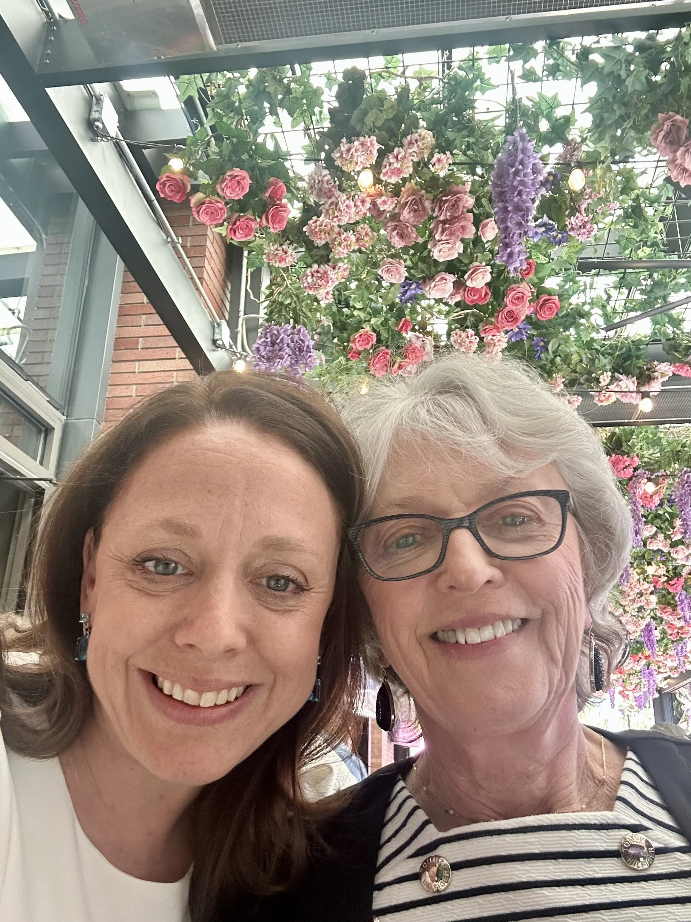 Two smiling women taking a selfie in front of an indoor floral display with pink, purple, and red flowers hanging from the ceiling.