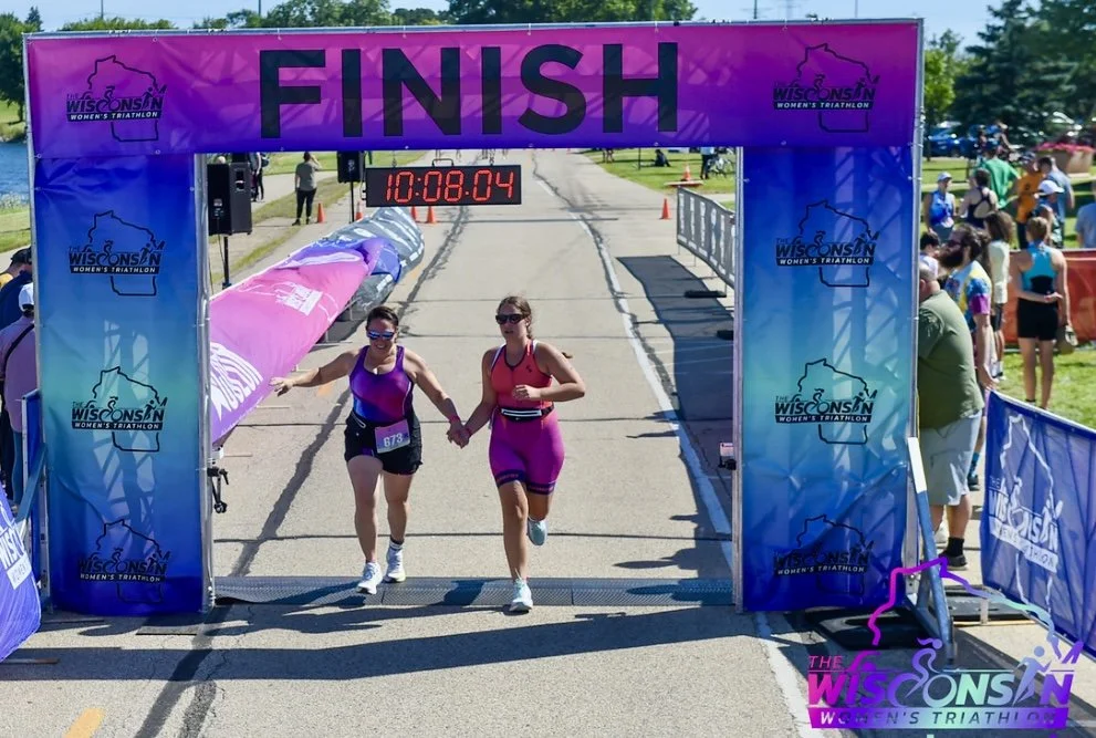 Two women finish a women's triathlon race holding hands under a purple finish line banner that says "FINISH." A digital timer showing 10:08:04 is above them. Spectators stand on the side watching.