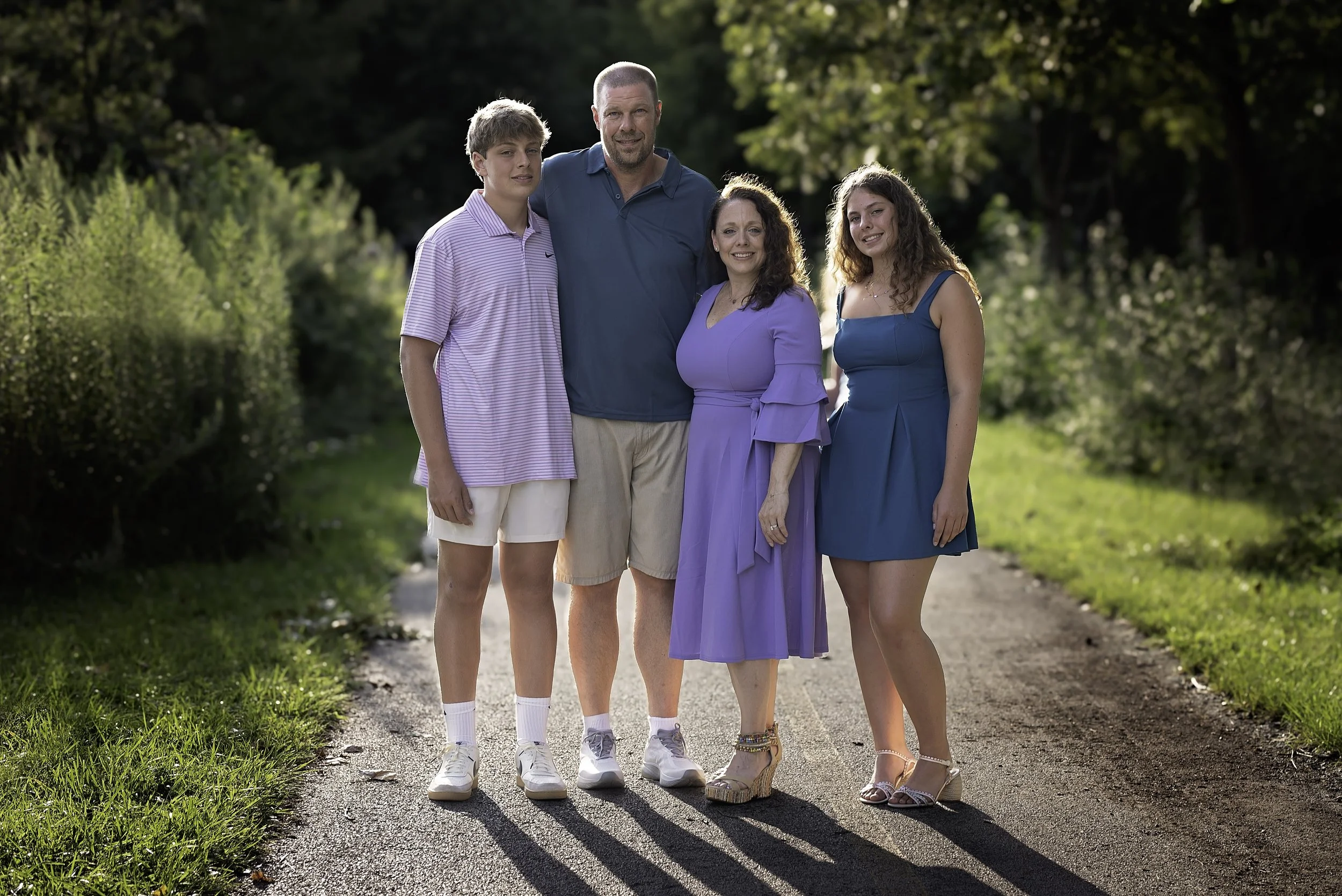 A family of five standing outdoors on a dirt path surrounded by greenery. The group includes two young adults, an older man, and two middle-aged women, all dressed in casual summer clothing.