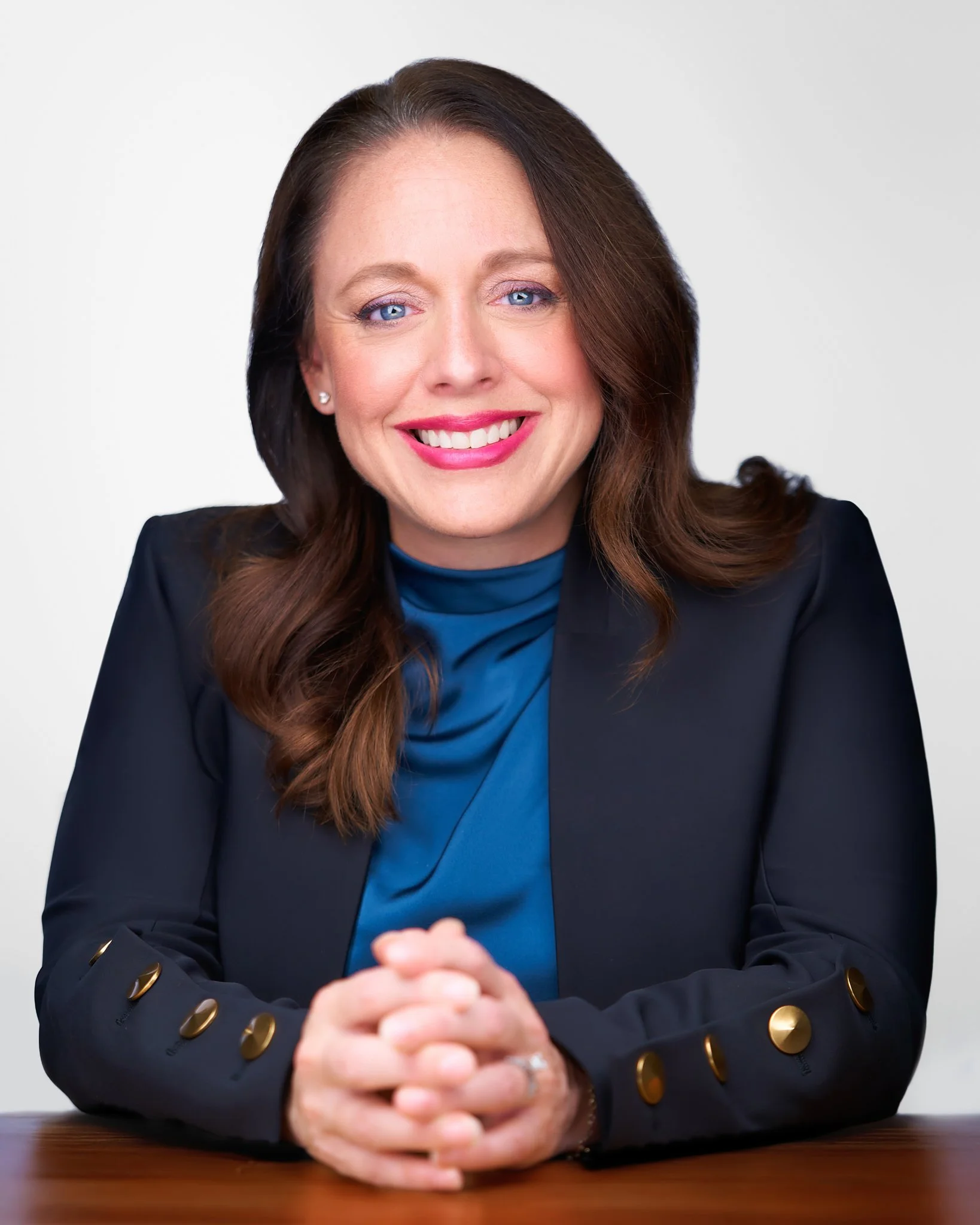Professional woman with long brown hair and blue eyes, smiling, wearing a black blazer with gold buttons and a blue shirt, sitting at a wooden table with hands clasped.