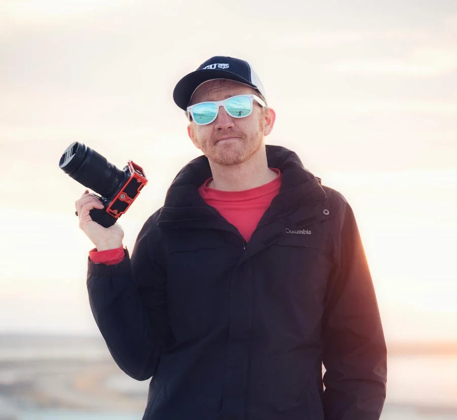 A man wearing a black Columbia jacket, red shirt, white sunglasses, and a black cap with sunglasses on, holding a camera with a large lens, standing outdoors at sunset or sunrise.