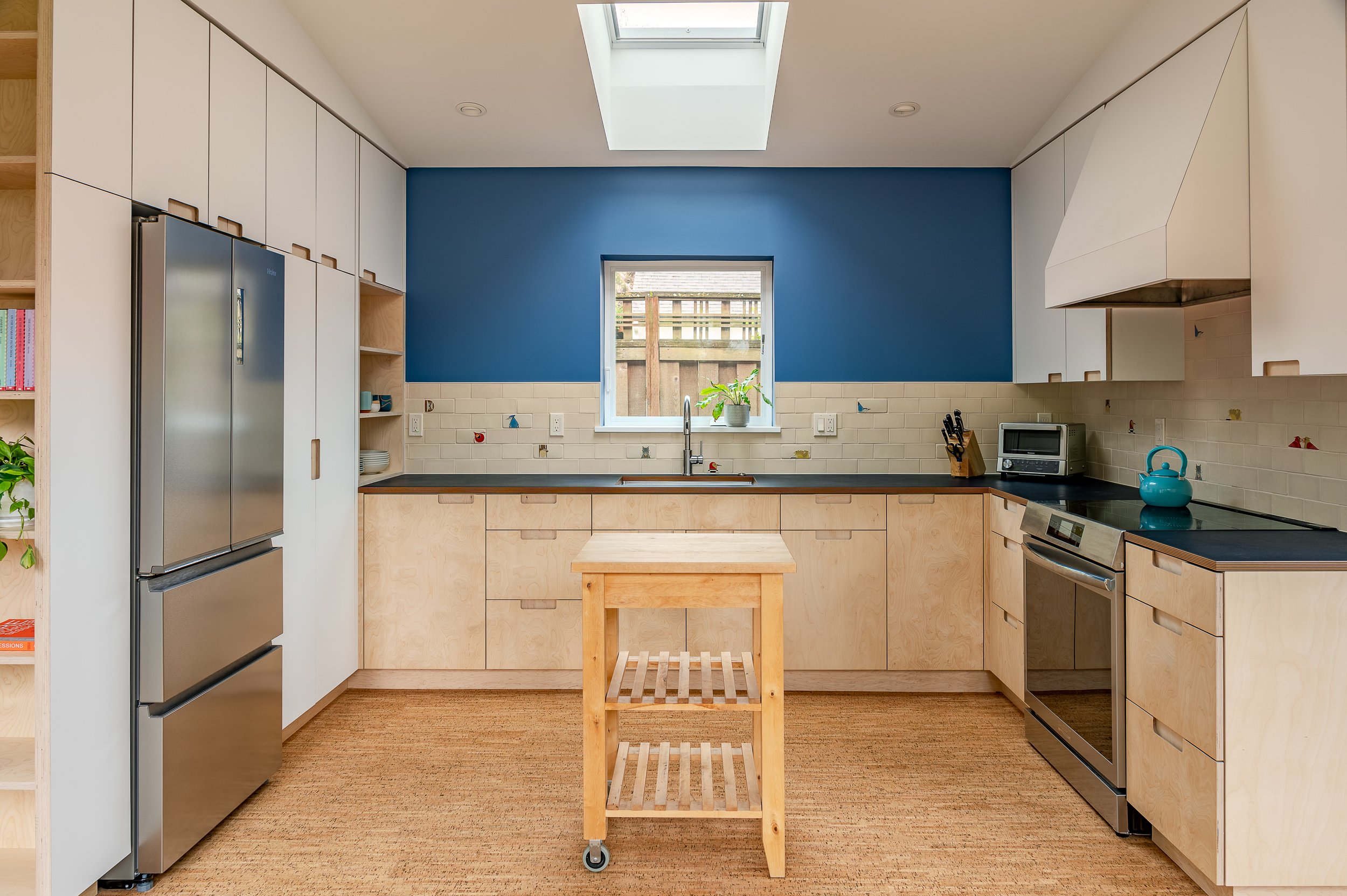 ADU kitchen with a blue accent wall, wooden cabinets, stainless steel refrigerator, microwave, teapot, and a small wooden island on wheels, illuminated by natural light from a skylight and small window.