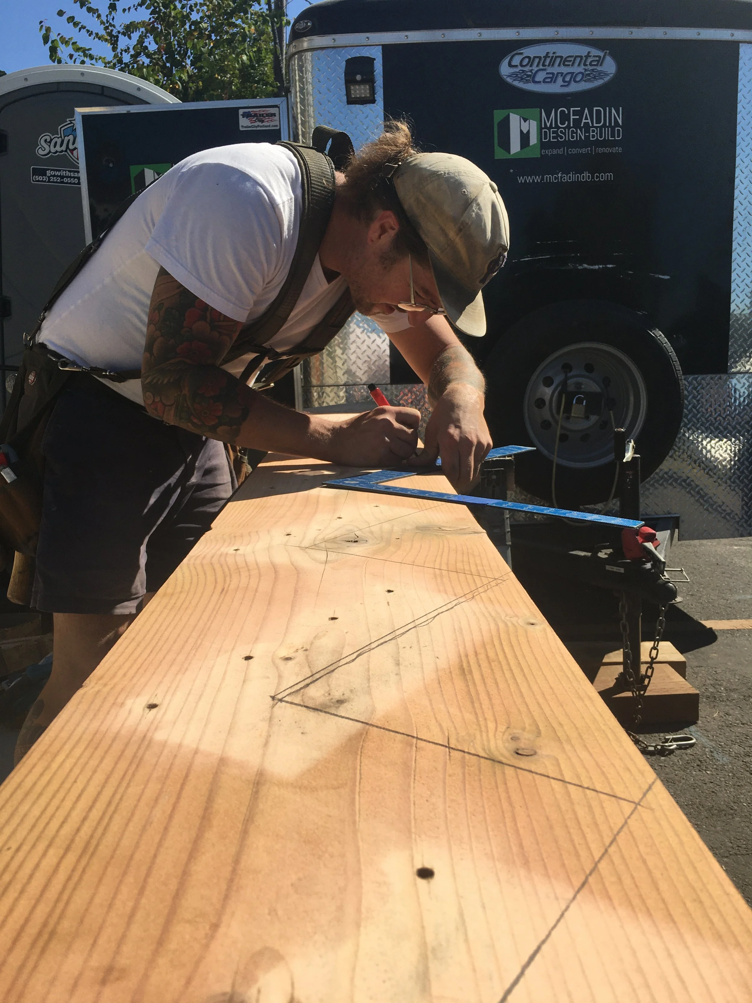 A man with tattoos on his arm, glasses, wearing a beige cap and white t-shirt, is working on a piece of wood outdoors, sketching with a pencil, with a trailer in the background.