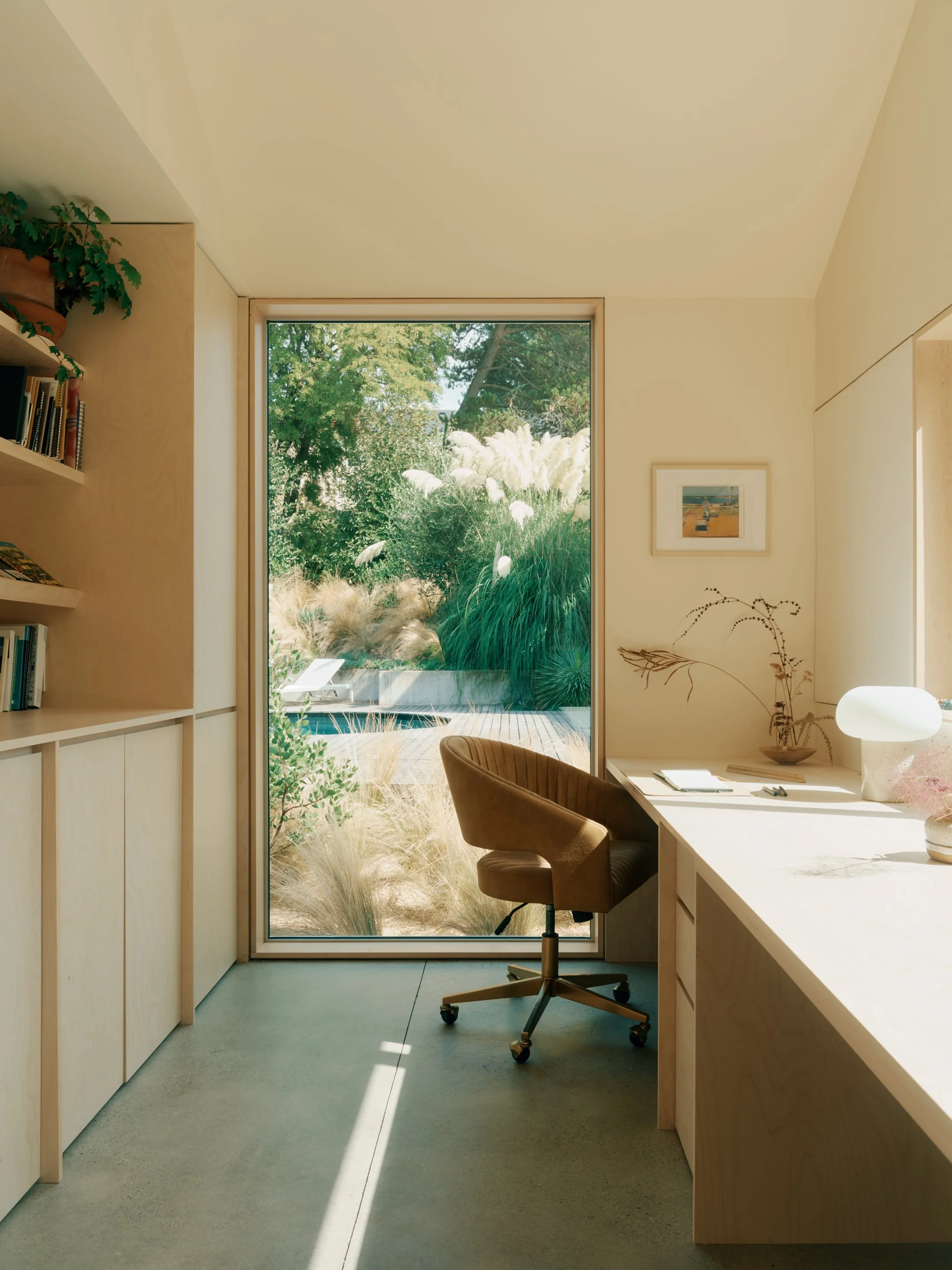 A home office in a pool house with a view of a lush outdoor garden through a large window. The room has light wood cabinetry, a beige office chair, and a white desk with decorative plants and a lamp.