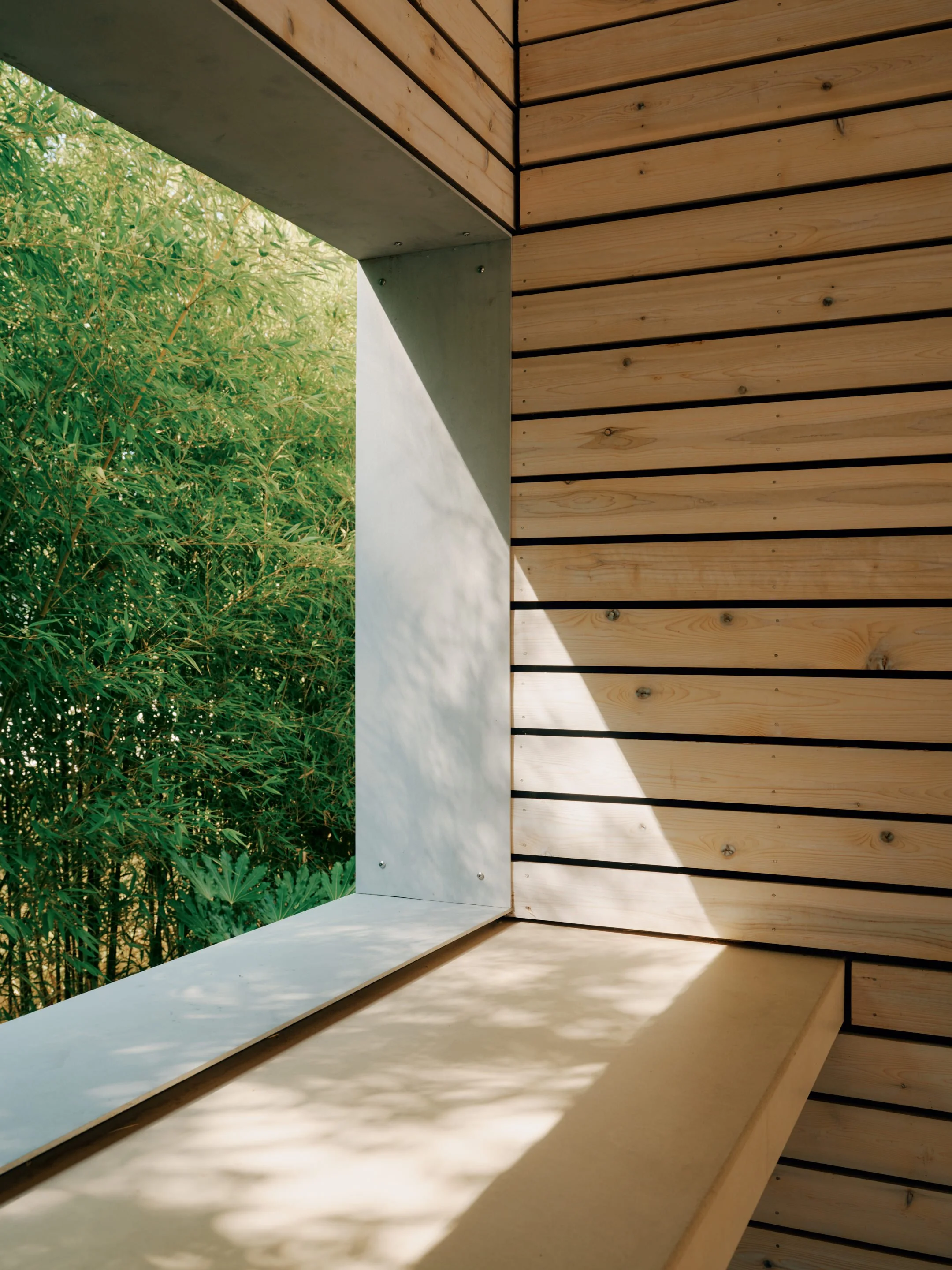 Part of a window with a white ledge and a wooden wall with horizontal planks in a pool house, overlooking green foliage and trees outside.