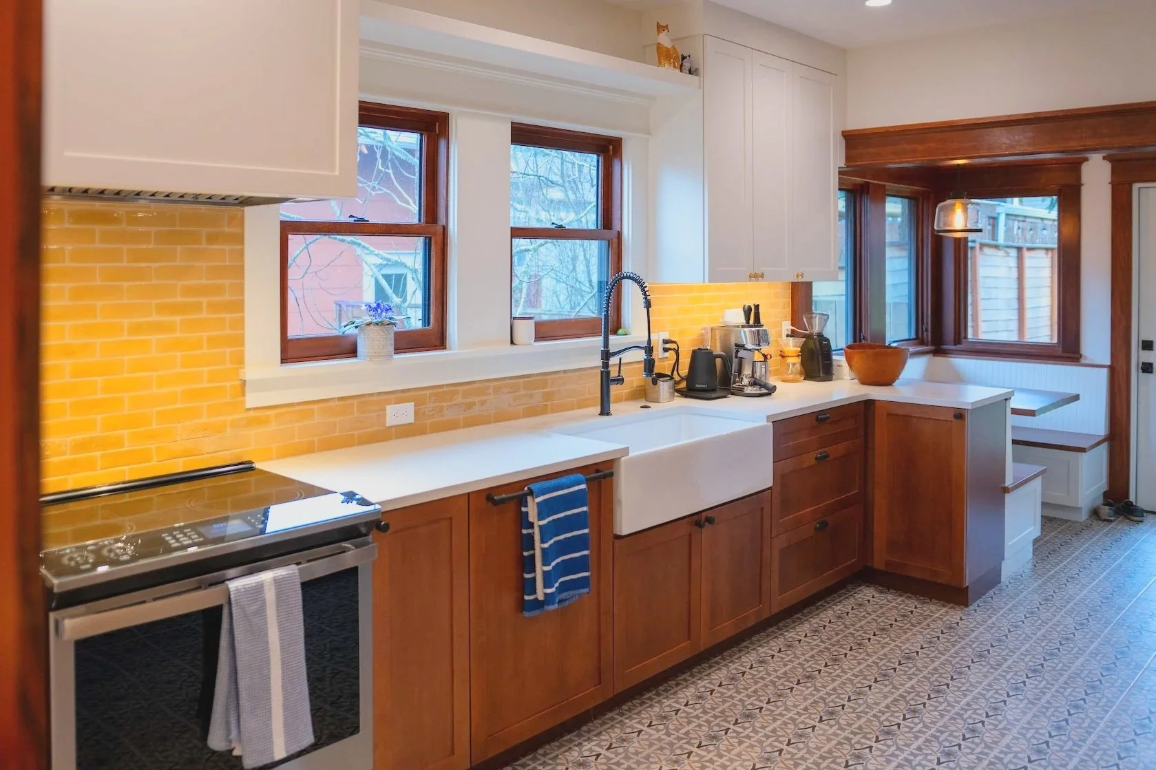 View of the remodeled counter with oven, white countertop, sink, coffee area, and banquette seating, rich dark wood, earth-toned yellow tile, dark faucets, and lots of windows to let in natural light.