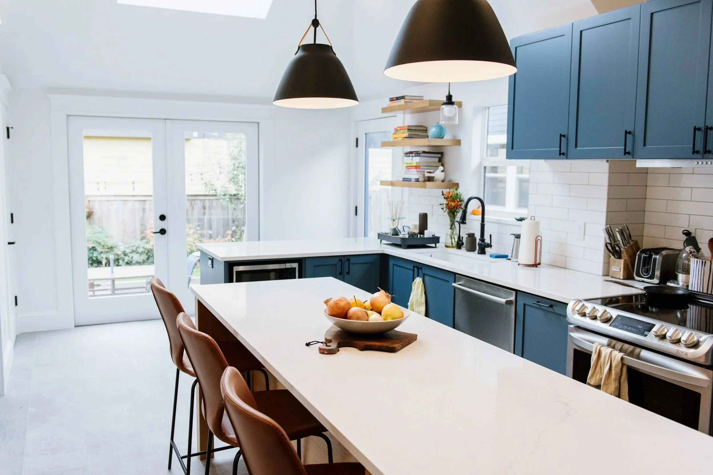 Modern kitchen with white island counter, blue cabinets, black pendant lights, and a bowl of fruit on the island.