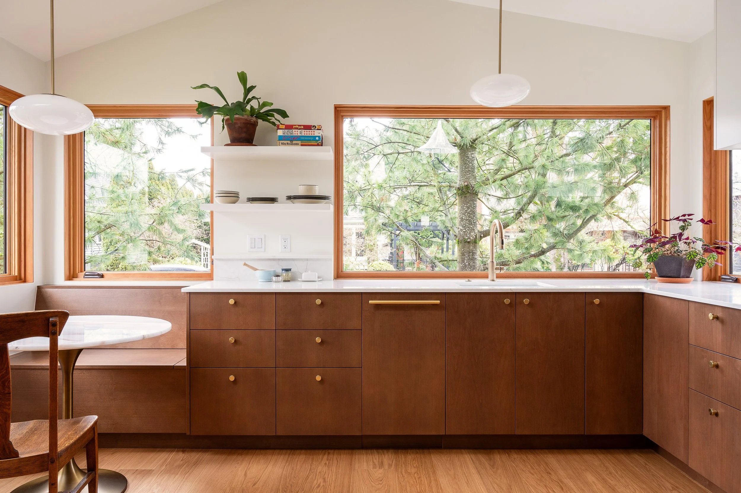 Kitchen with large windows showing trees outside, wooden cabinets, and a white countertop. There are plants on windowsills, and open shelving with dishes and books.