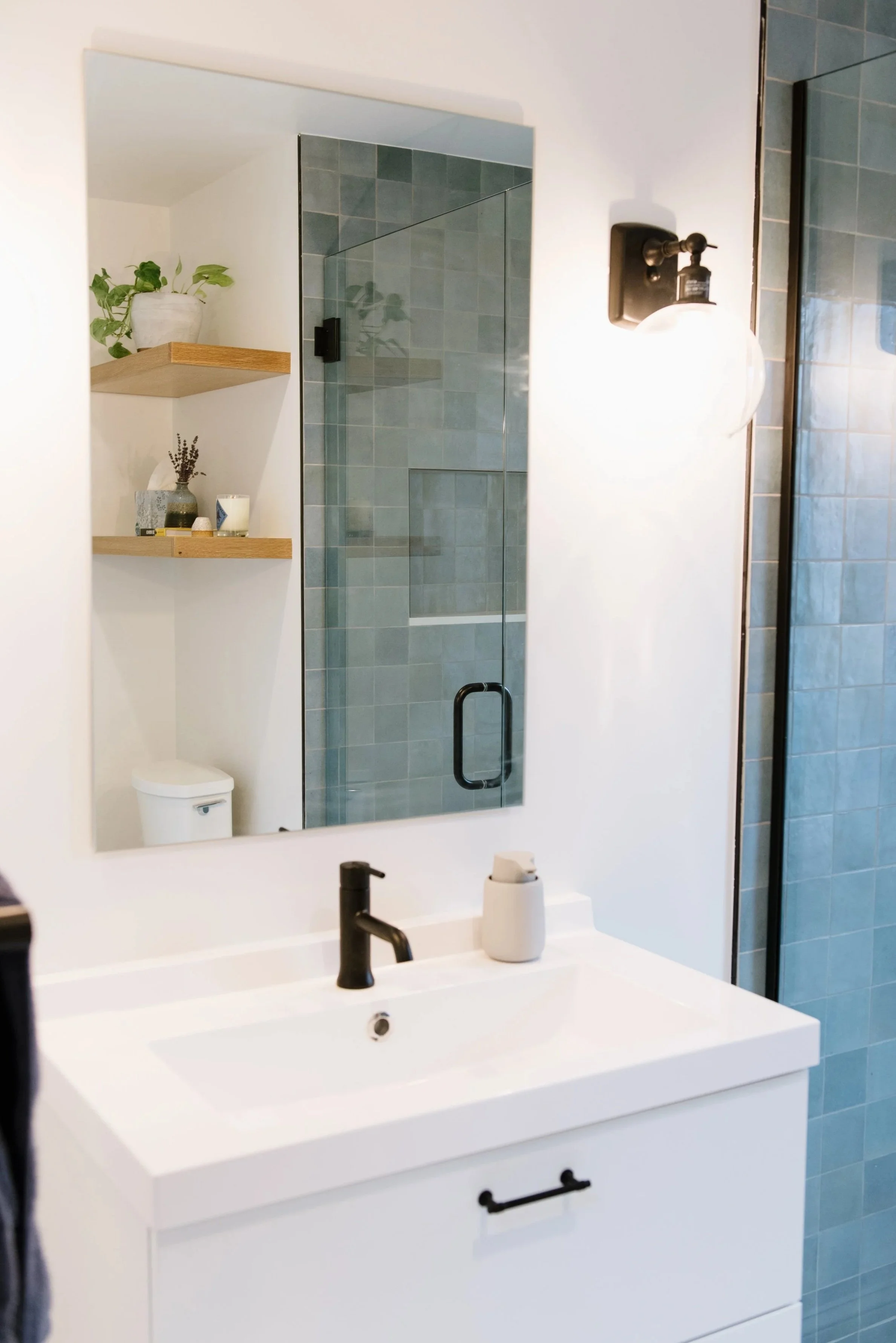 White bathroom with dark metal faucets and fixtures, large, rectangle mirror above, and green tiled shower with clear glass door.