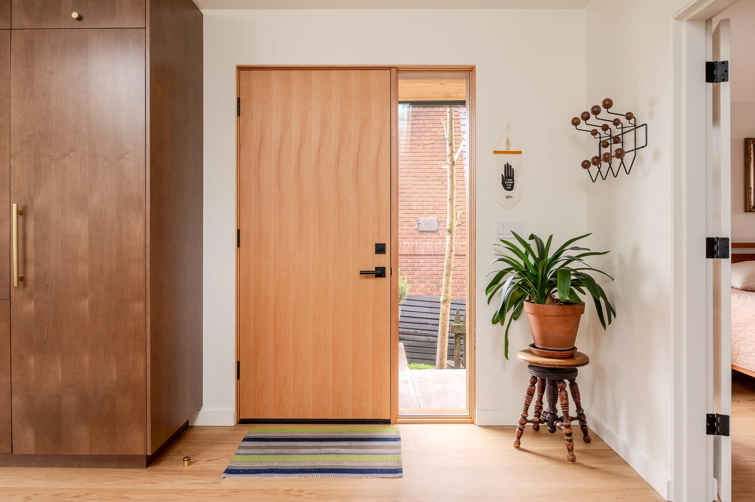 Interior view of an ADU entryway showing a wooden front door with side window, a potted plant on stool, wall decor, and part of a bedroom.