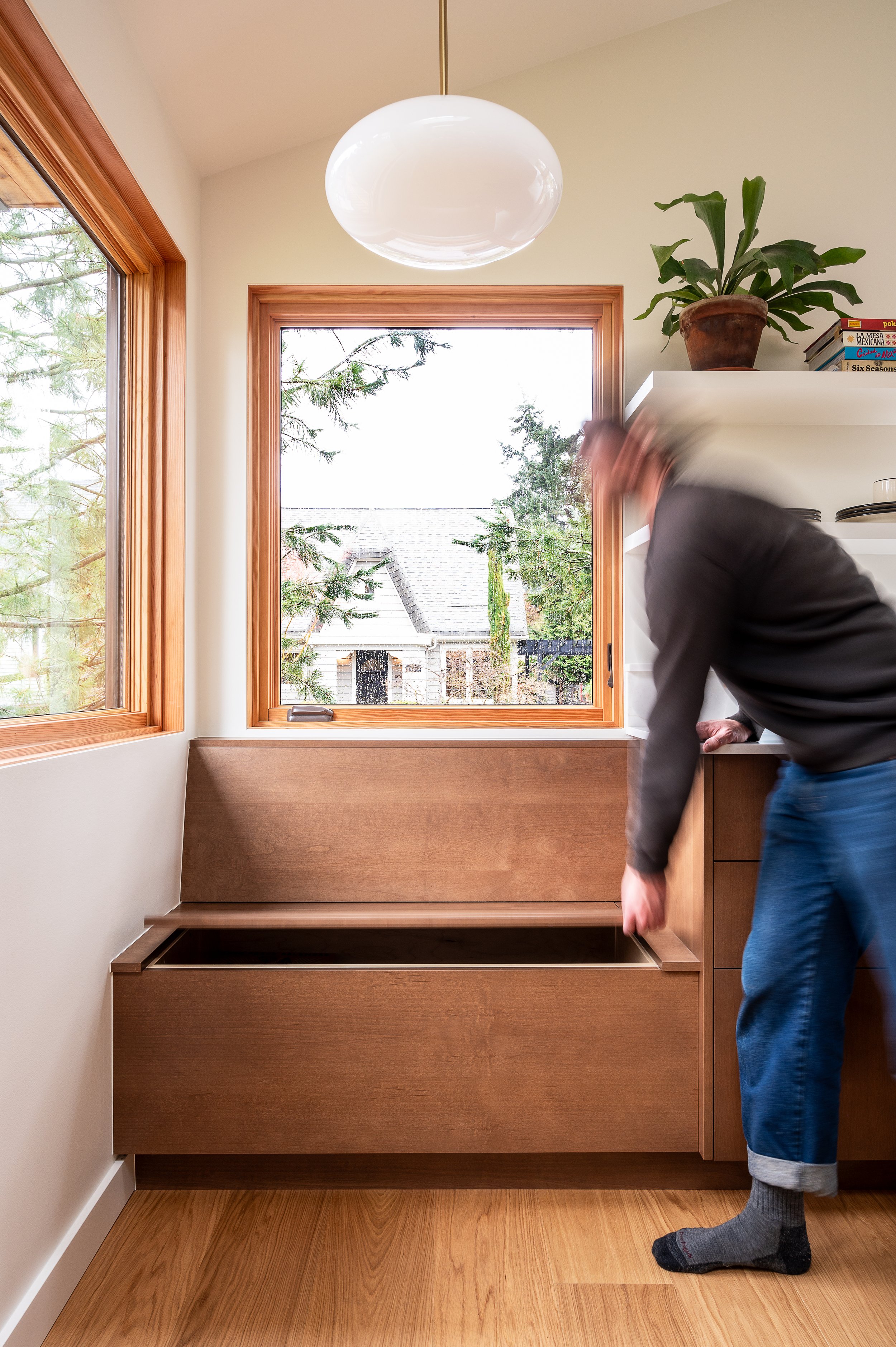  Modern ADU kitchen with large windows, wooden trim, a white ceiling, and wooden flooring, with a large built-in storage bench under the window and a person bending over to open the bench seat compartment.