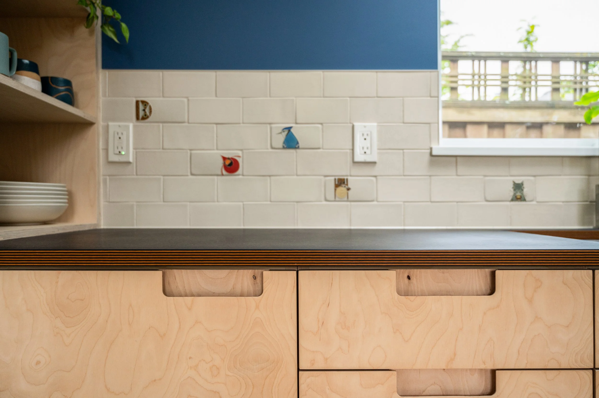 ADU kitchen wall with white subway tiles, decorated with small eco-themed stickers. To the left is a wooden shelf holding cups and bowls, and next to it are electrical outlets. A window on the right shows a wooden fence and some greenery outside.