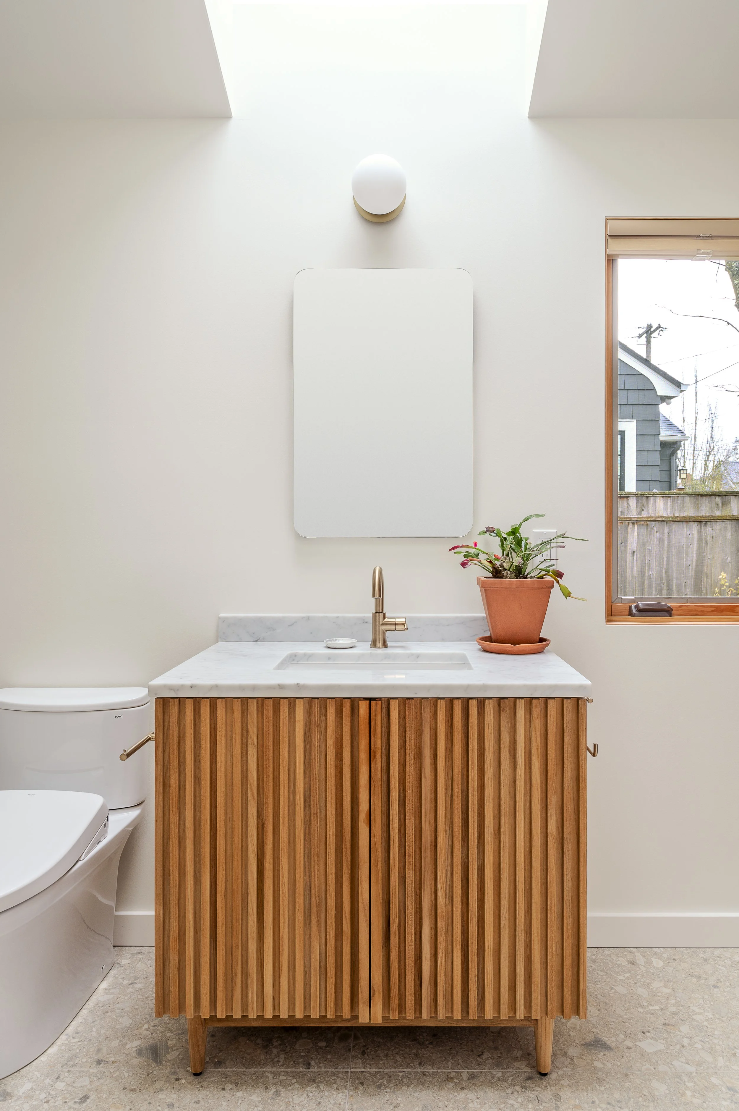 A modern ADU bathroom vanity with a white marble countertop, wood slatted cabinet, copper faucet, a potted plant, a mirror, and a wall-mounted light fixture next to a window.