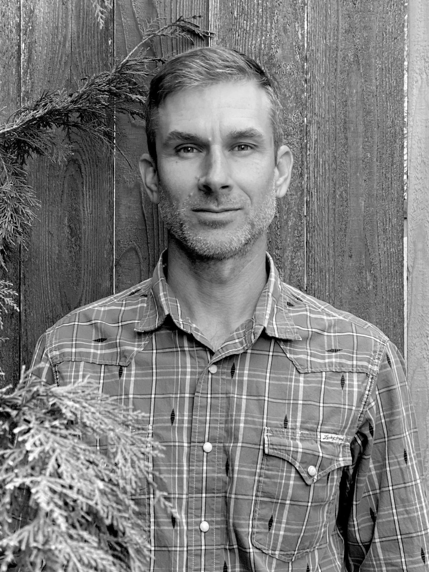 A black and white portrait of owner, Zeke McFadin, man with short hair, a light beard, wearing a plaid shirt, standing outdoors in front of a wooden fence with some pine branches in the foreground.