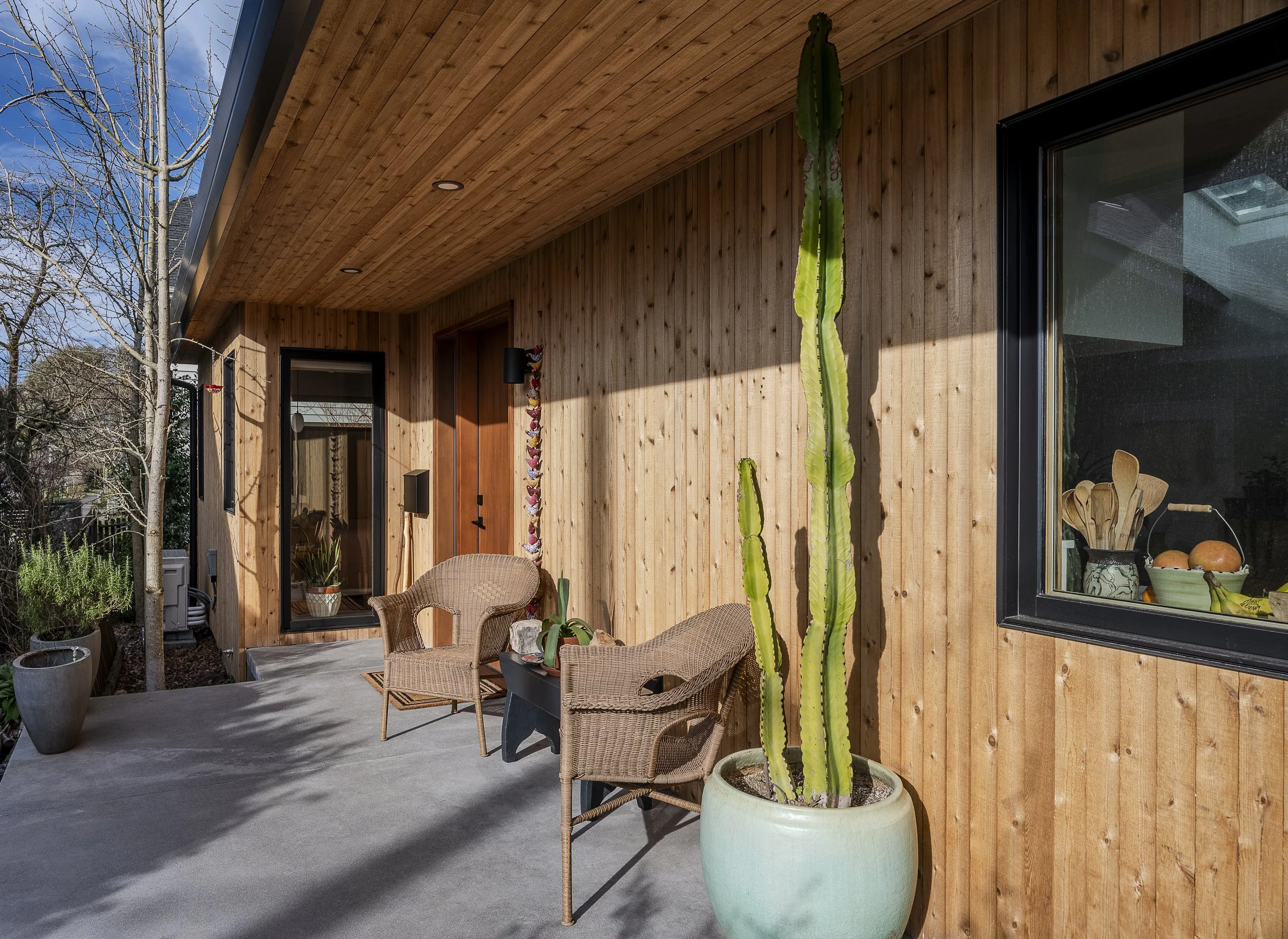 A new ADU porch with woven chairs, large potted cactus, and other potted plants. The house has a large window and a small decorative hanging beside the door.
