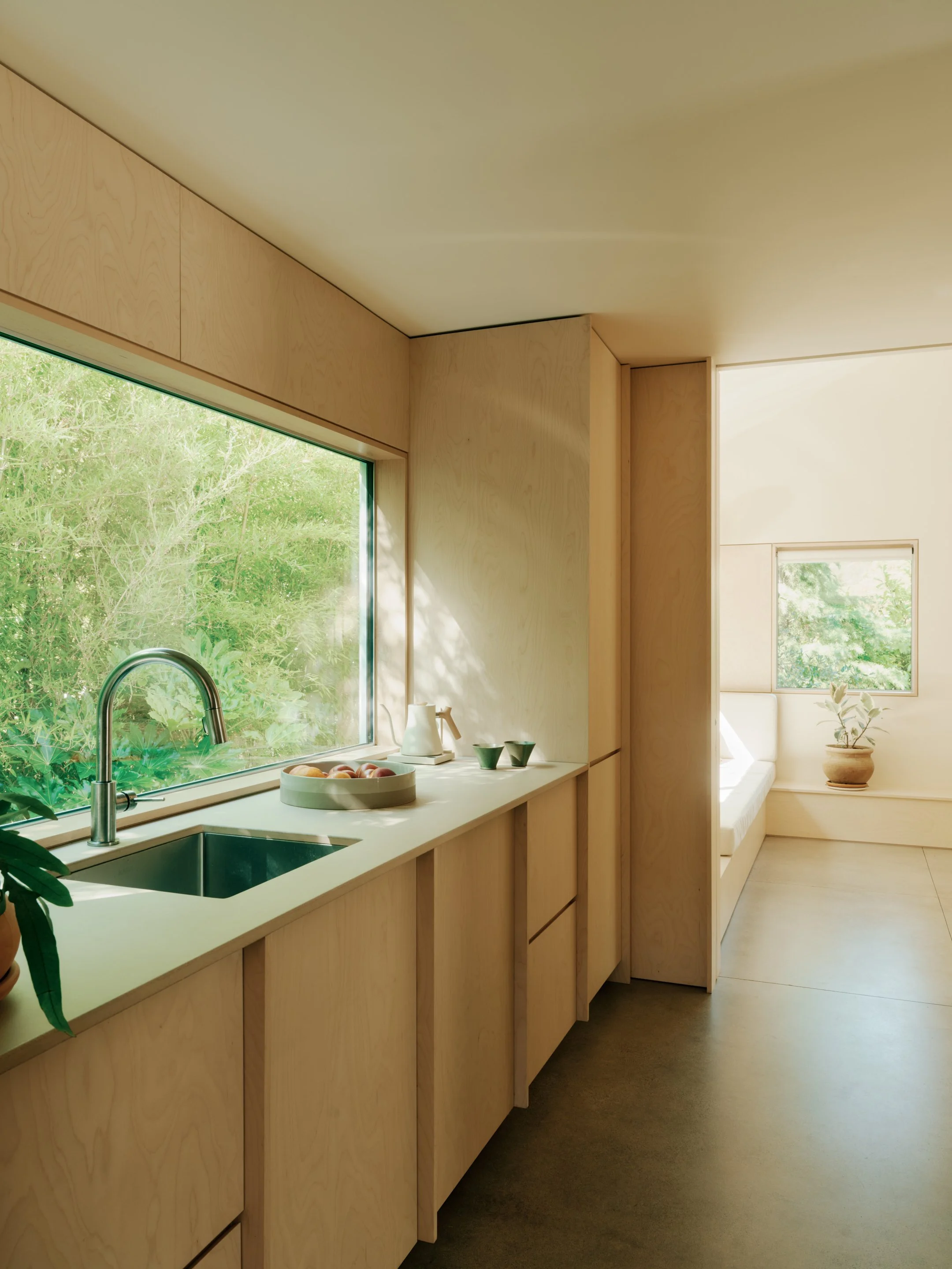 Modern pool house kitchen with light wood cabinetry, a large window with a view of greenery, and a seating nook with a potted plant.