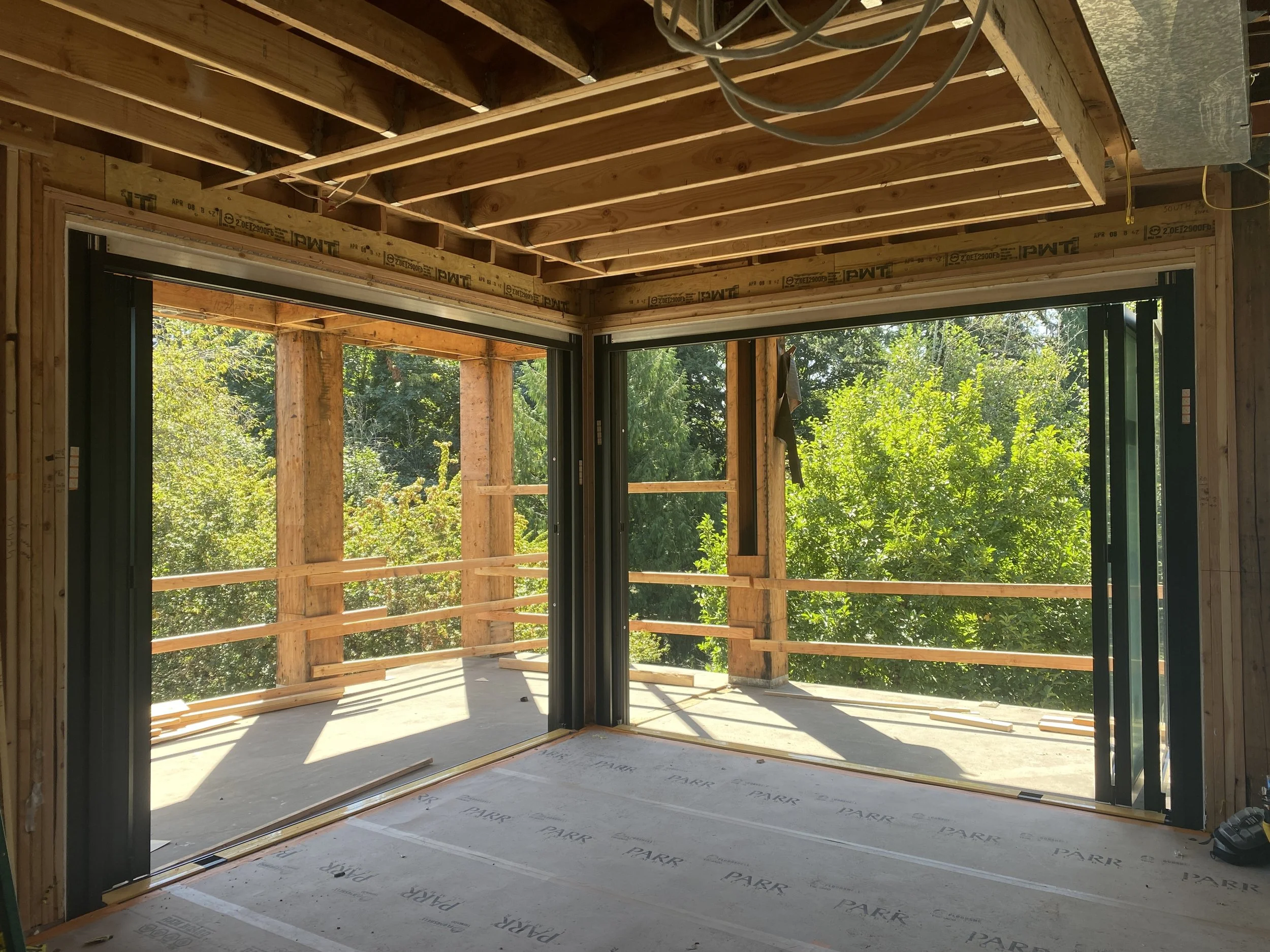 Interior of a house under construction with exposed framing, large sliding glass doors, and a view of a wooded outdoor area.