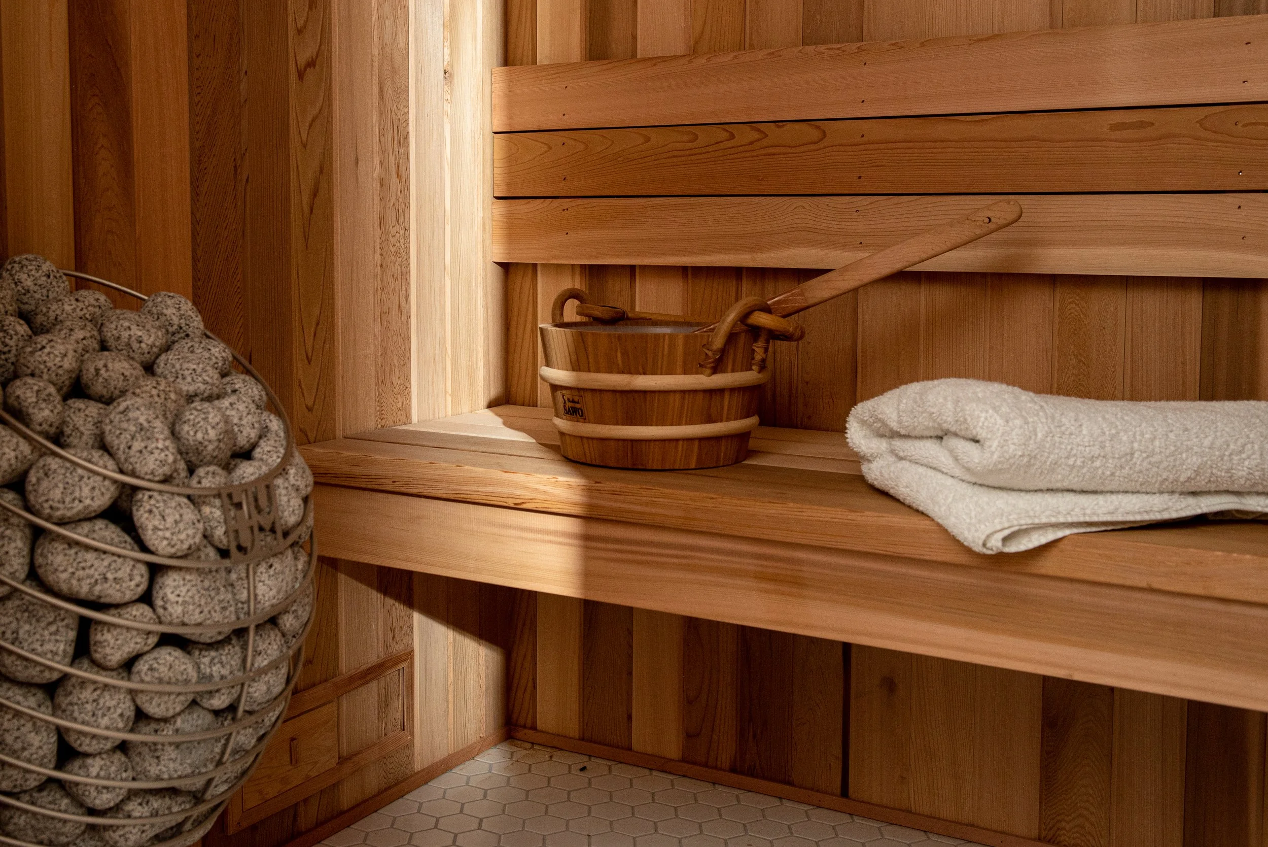 The inside of the cedar sauna addition to this basement remodel contains a seating bench.  It's holding a cedar water bucket with large wooden spoon a pile of white towels, and to the left, a HUUM heater with a metal cage holding smooth grey  stones.