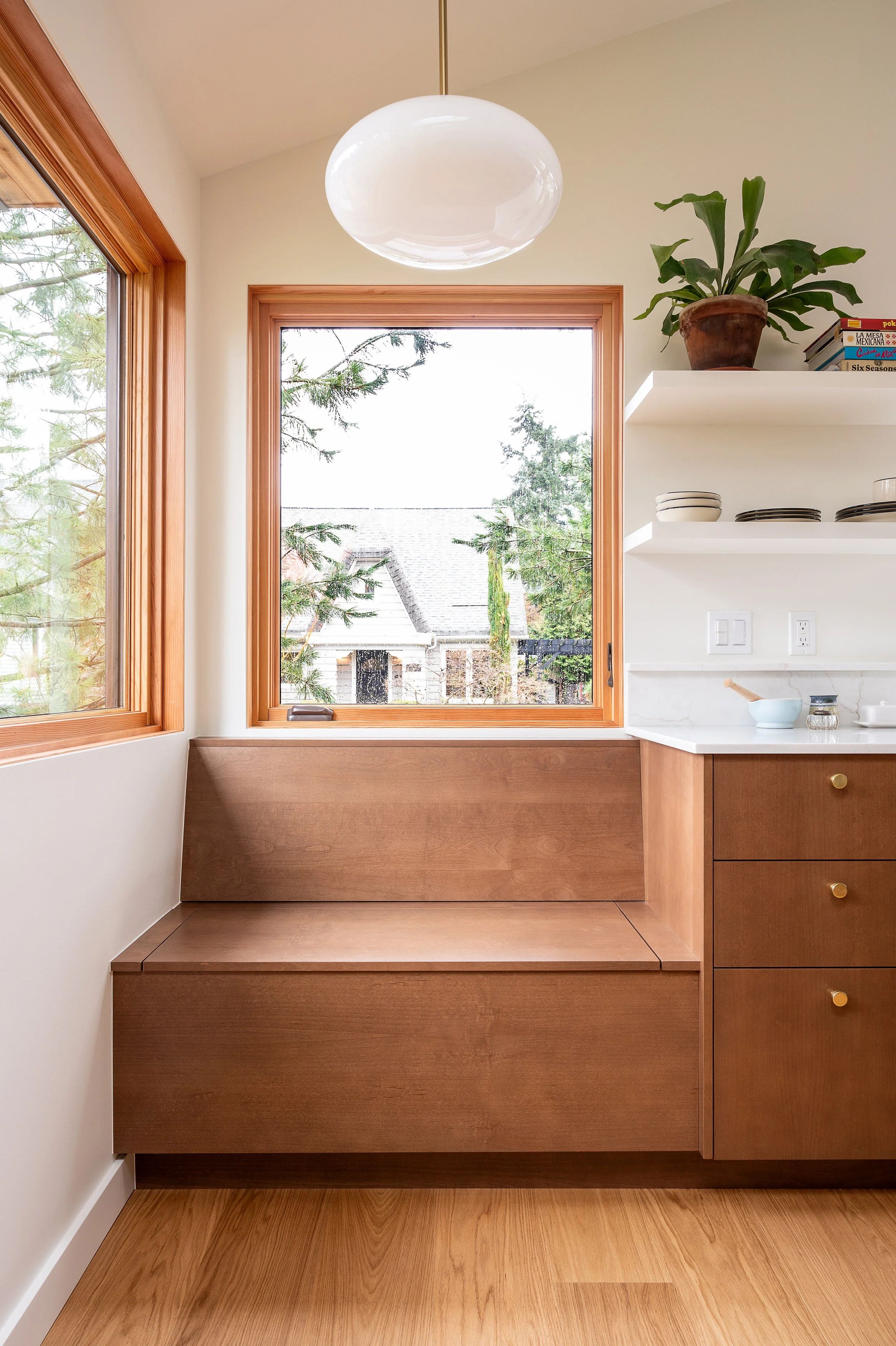 A cozy interior corner in a modern ADU with large wooden window frames, a built-in wooden bench, white walls, a white floating shelf with a potted plant and stacked 
dishes.
