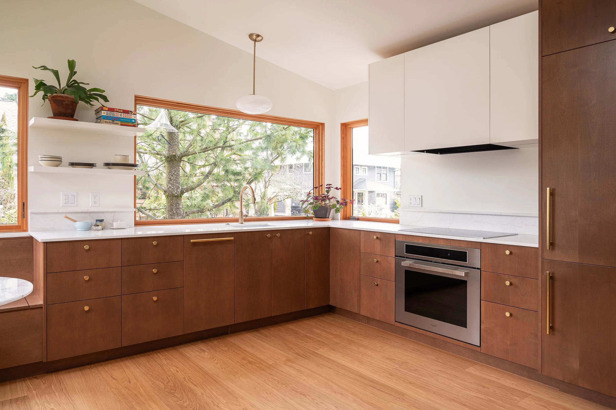 Modern and compact ADU kitchen with wooden cabinets, white countertops, large corner window, small open shelf with plates and bowls, potted plant, electrical outlets, and built-in oven.