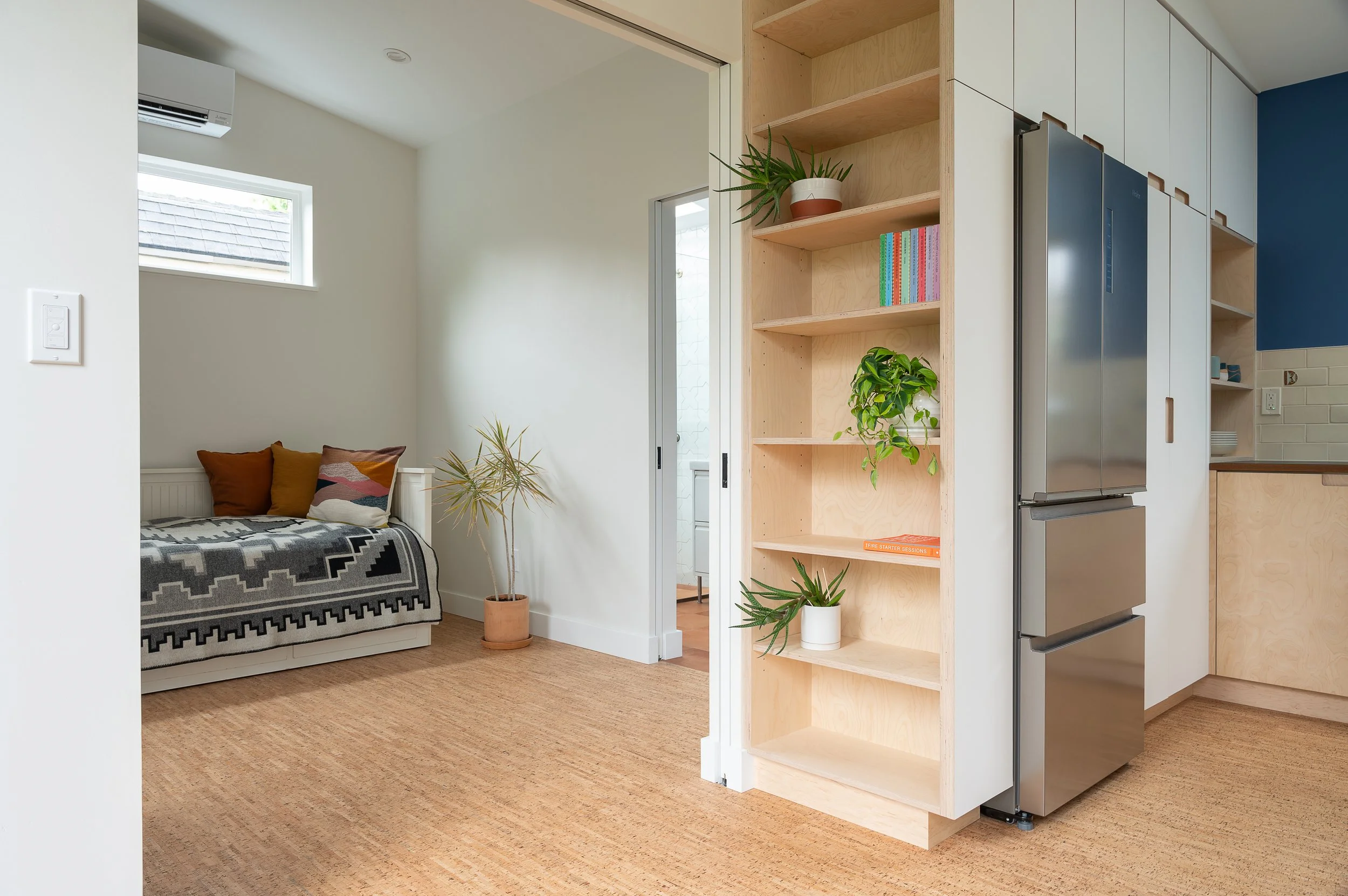 Interior of ADU with a view of a bedroom and kitchen area, featuring a bed with colorful pillows, a potted plant, wooden shelves, and a stainless steel refrigerator.