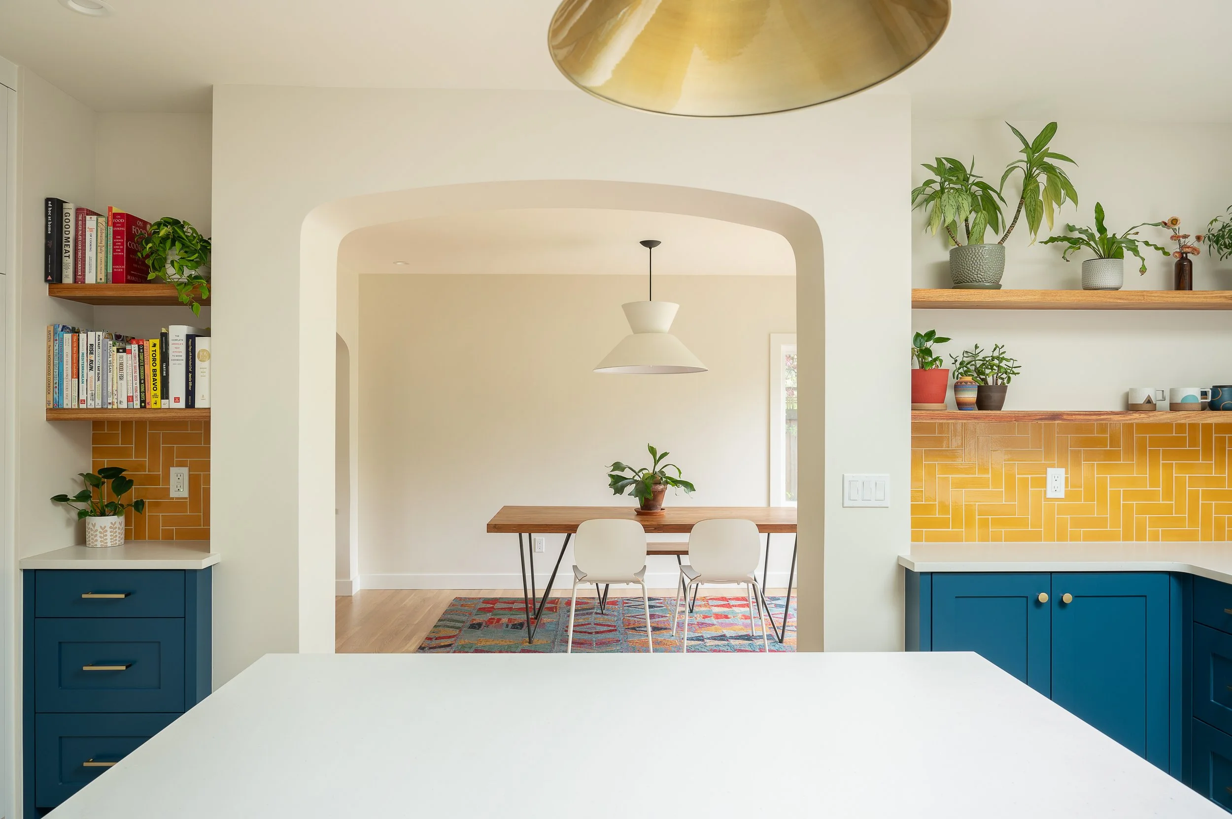  Remodeled dining area and kitchen. The walls are painted cream, and there are wooden shelves with plants and books. There are two pendant lights hanging from the ceiling, and the space is well-lit with natural light.