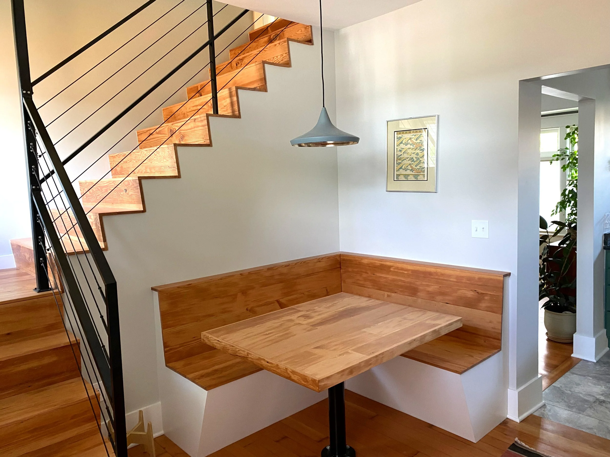 Interior of a modern home featuring a staircase with wooden steps and a black metal railing, a contemporary gray pendant light hanging from the ceiling, a framed artwork on the wall, a wooden bench built into the wall, and potted plants near the door