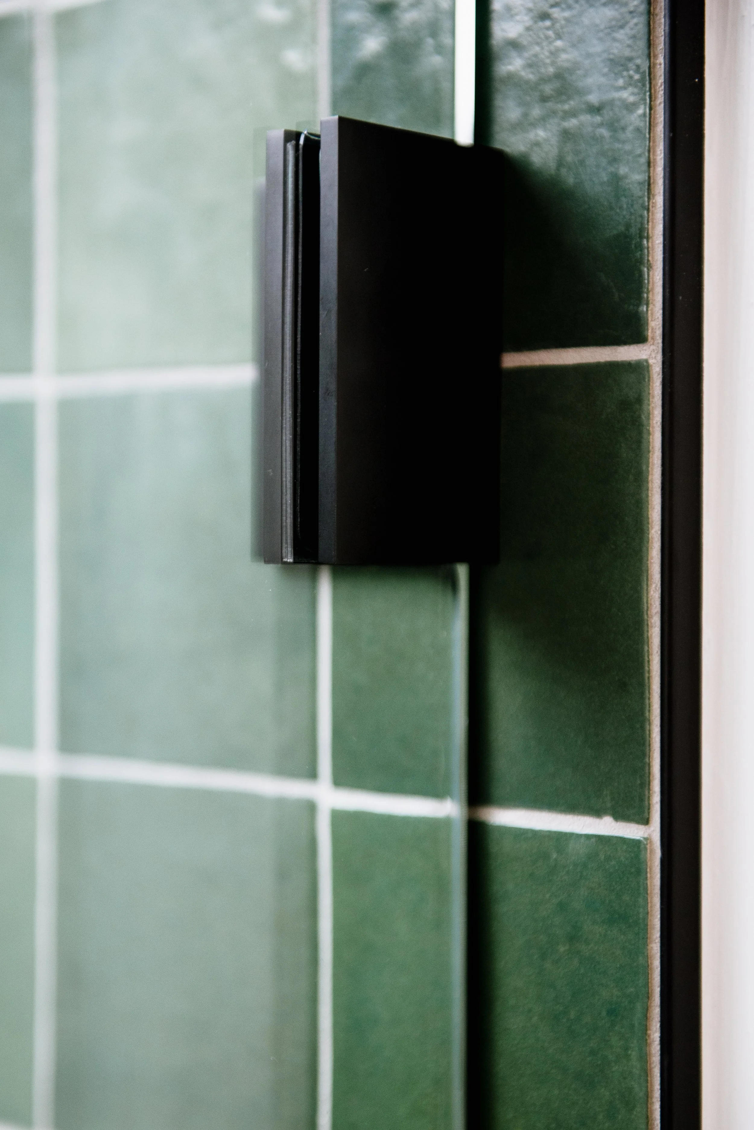 Remodeled bathroom with lose-up of a black door handle on a clear glass shower door with green tiled shower walls in the background.