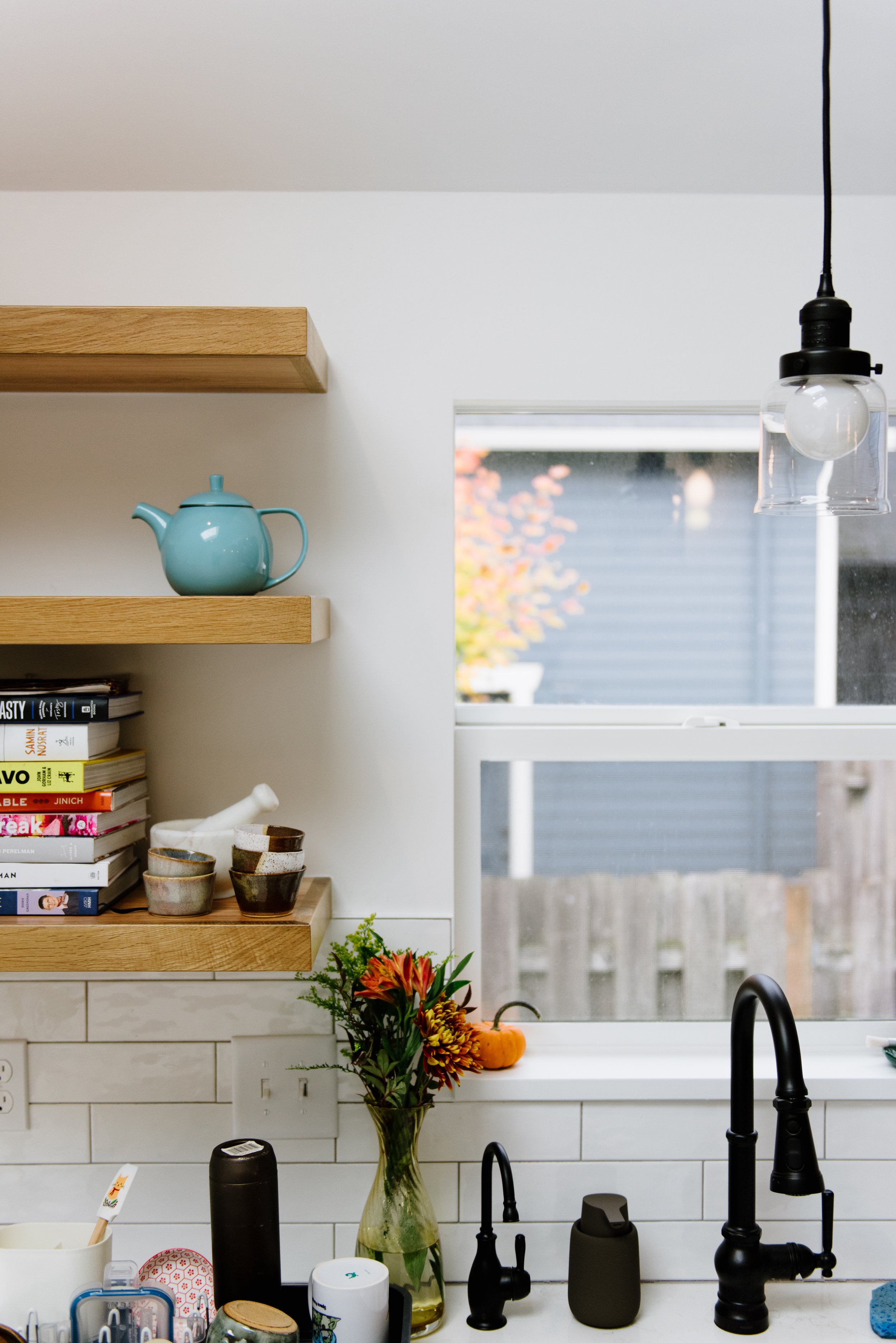 This renovated kitchen has with walls and trim around the window above the sink, dark metal faucets, and wooden floating shelves above the counter to the left.