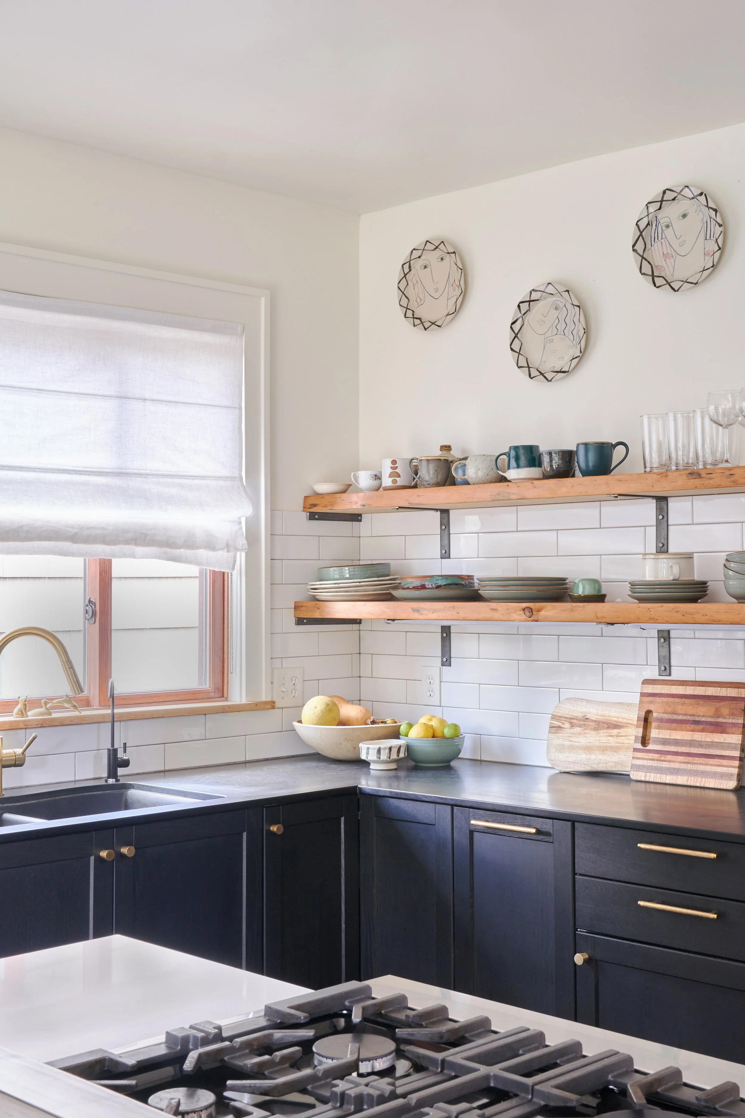 The corner of this renovated kitchen has white walls and tile, rustic wood shelves, dark grey cabinets, and an island with gas stove top.