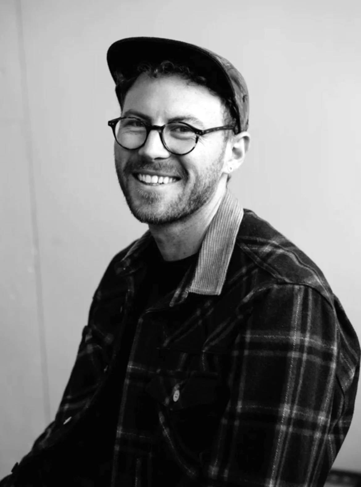 A smiling man with glasses, a beard, and a cap, wearing a plaid shirt.