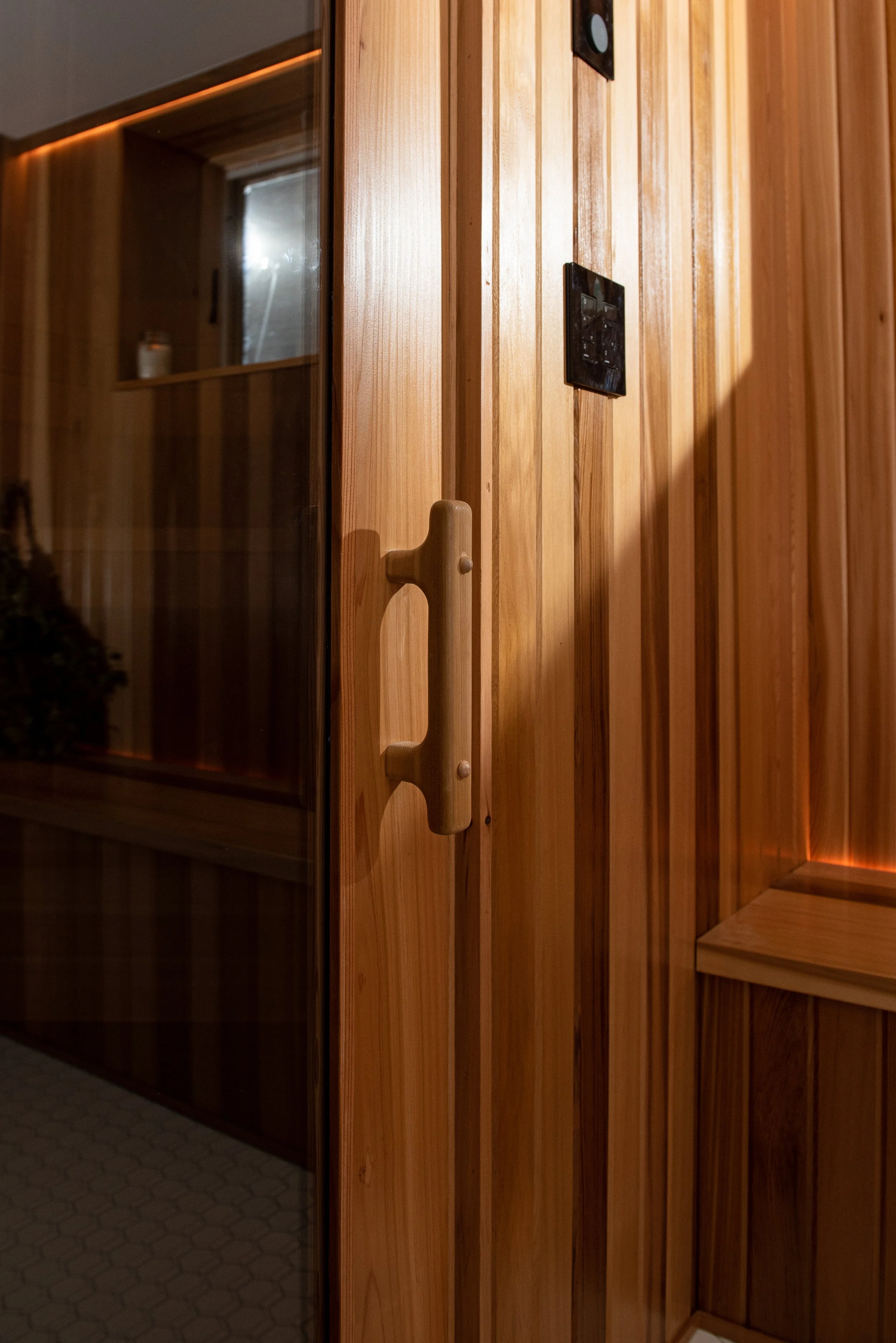 Outside the glass door of the cedar sauna basement renovation is a cedar bench, with rectangular widow above, and unobtrusive yet helpful light coming from the crack where bench meets wall.