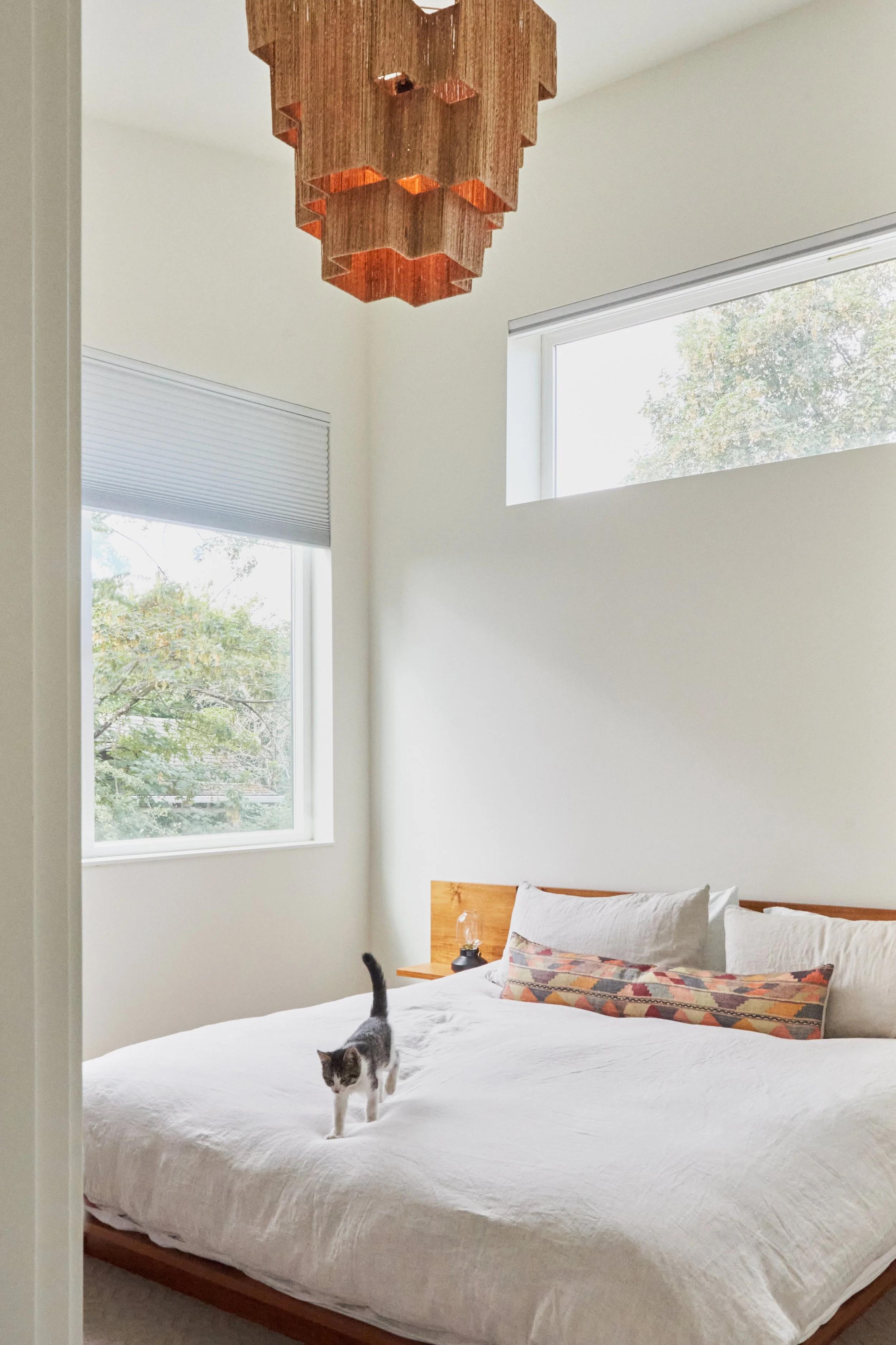This remodeled bedroom has 2 large tree-filled windows providing abundant natural light, white walls, and a modern geometric light fixture hanging over the bed. 