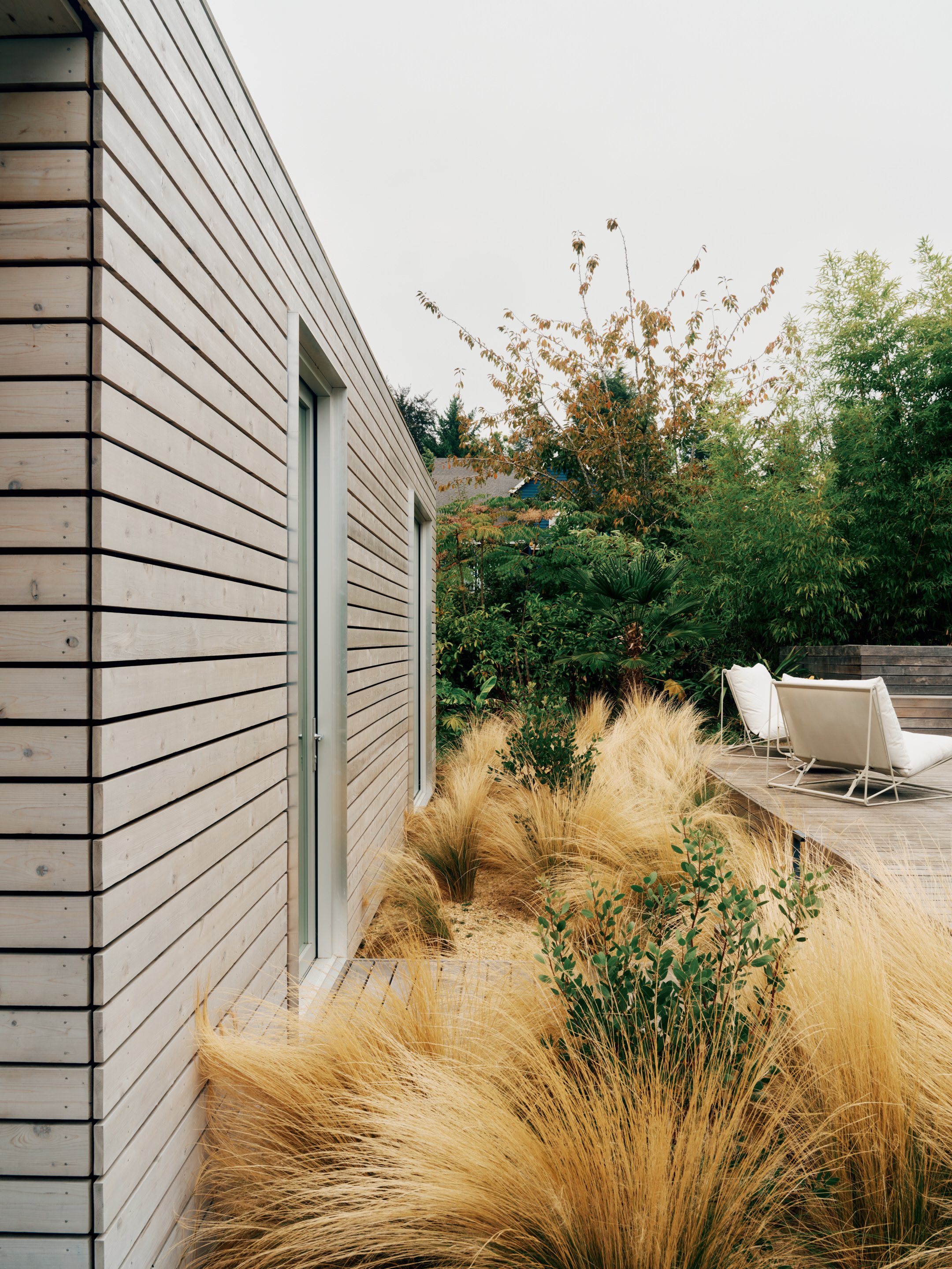 Outdoor patio with a modern pool house wall made of horizontal cedar planks, surrounded by tall ornamental grasses and lush green trees, with a seating area featuring white chairs and cushions.
