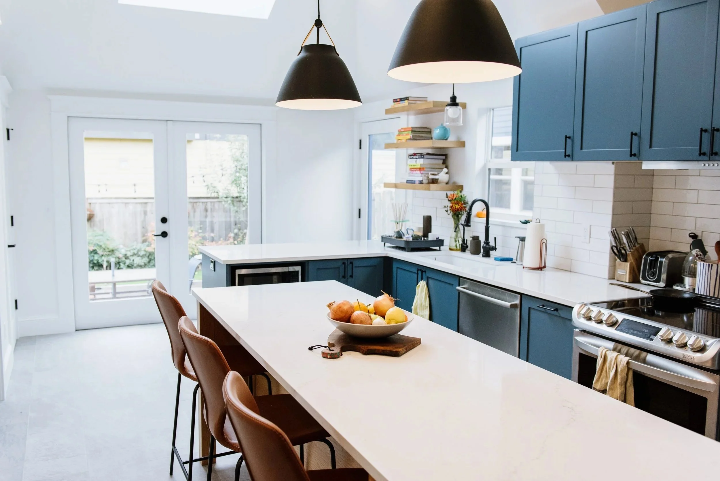 This remodeled kitchen has white floors, walls, and countertops, rich blue cabinet faces, dark metallic fixtures, and tanned leather bar stools at a seating area on the large kitchen island.