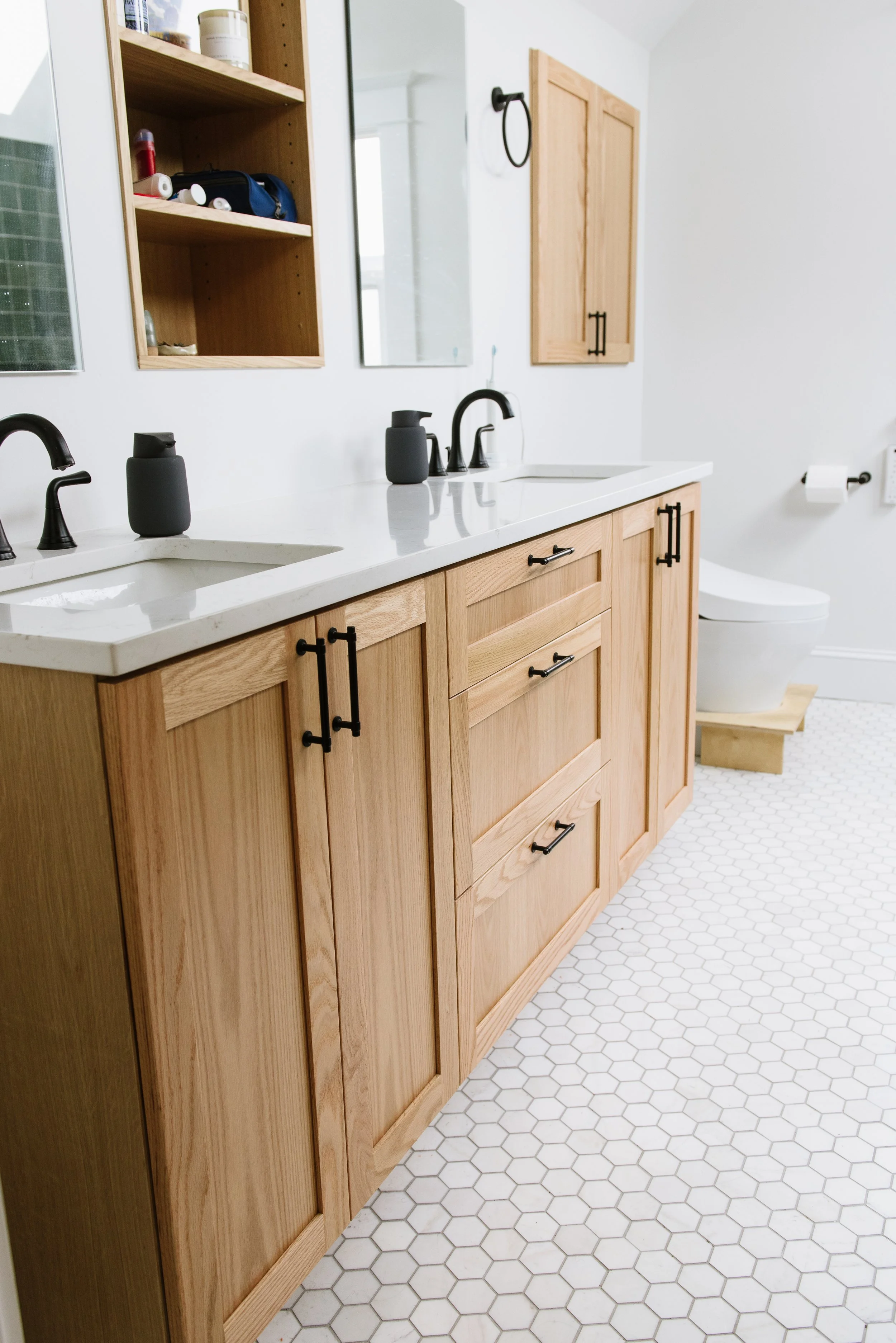 This remodeled primary bathroom has a double sink, finished wood cabinets, white walls, counters, and tile floors. A built in shelf in the wall between the sinks adds a nice vertical space for your things.