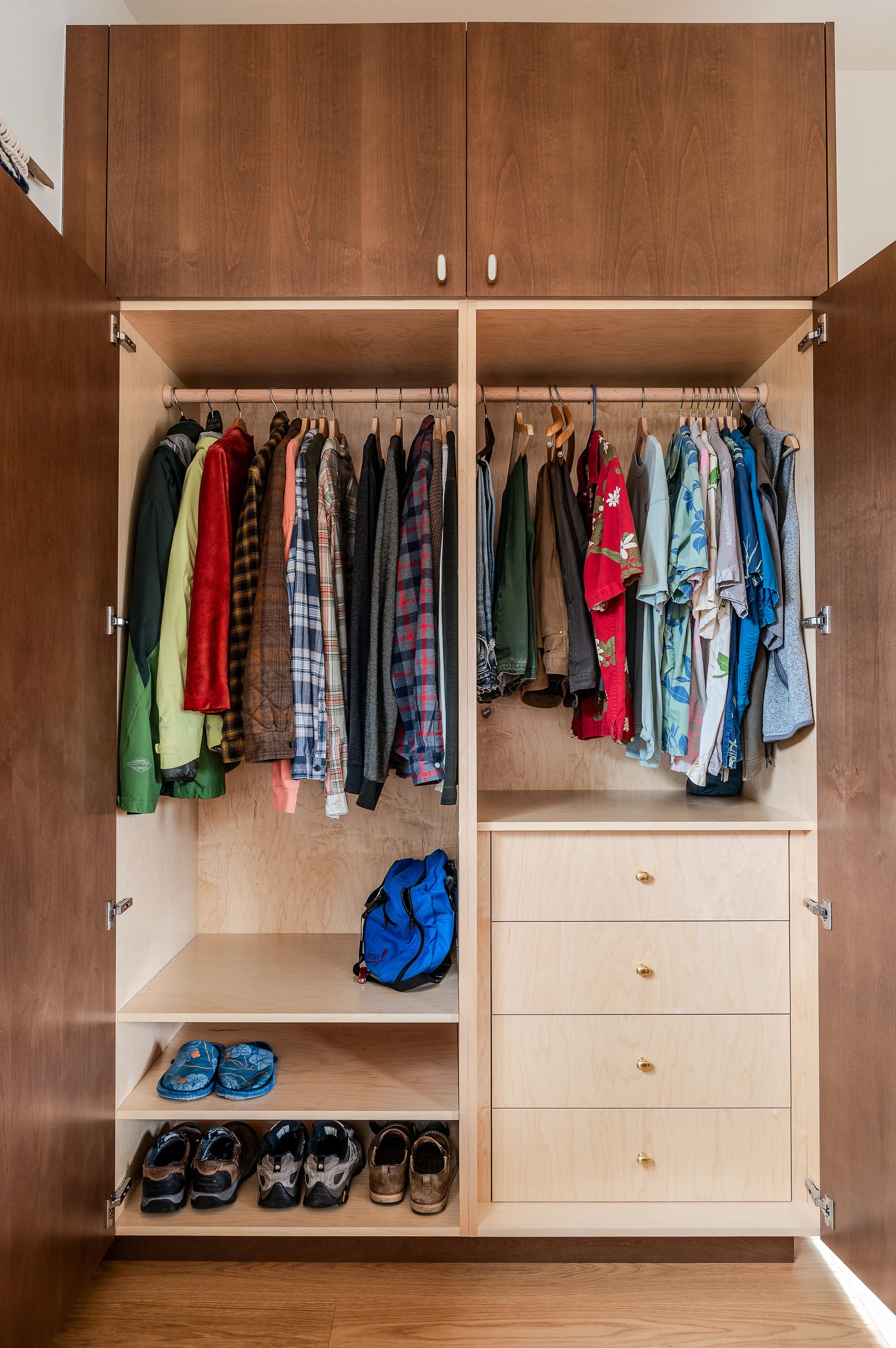 Open wooden closet wardrobe with hanging shirts, jackets, and shorts, along with shoes, sandals, and a blue backpack inside.