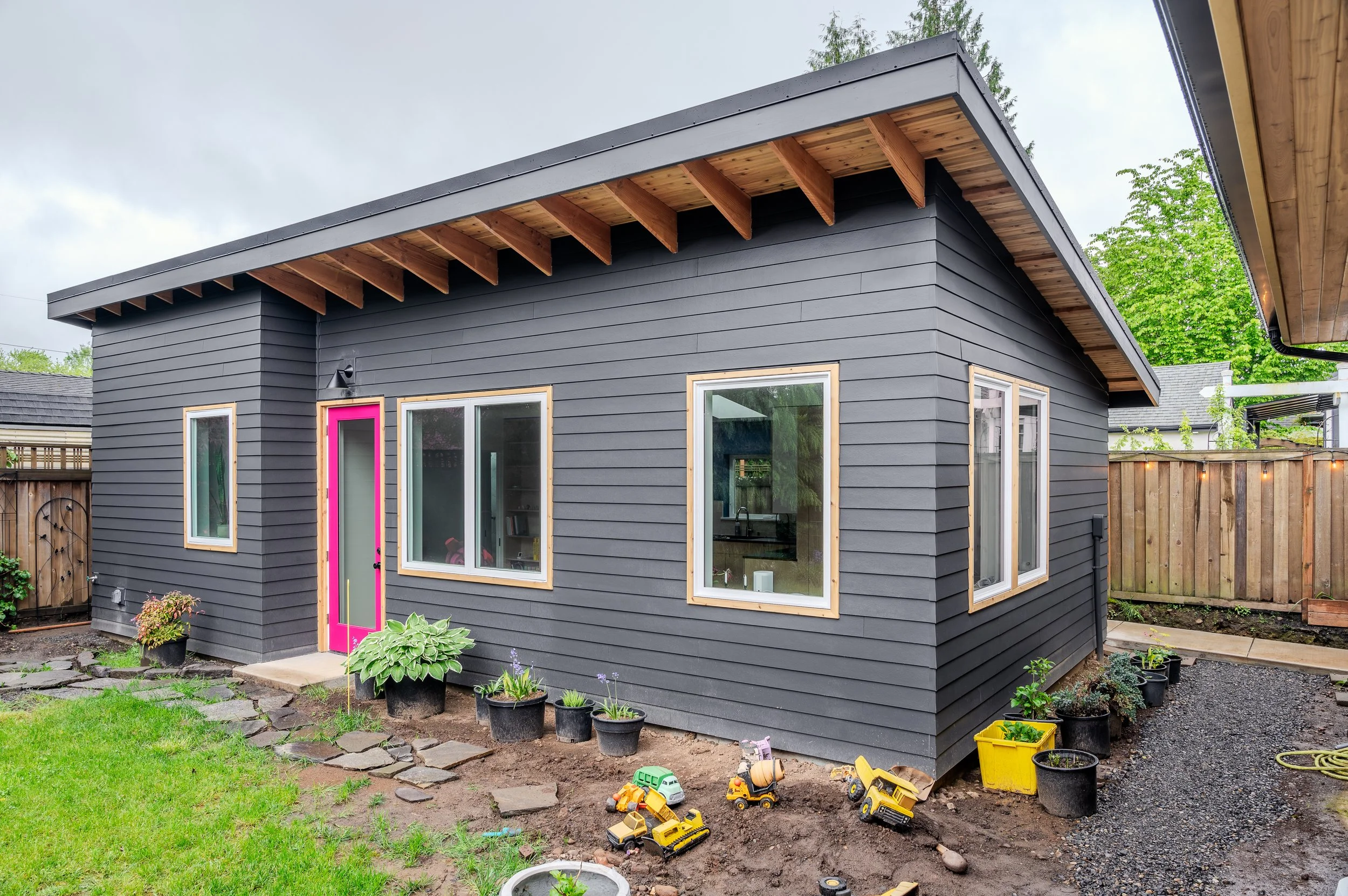 ADU for family to age in ploace with dark gray wooden siding, large windows, and a pink door. There are potted plants and gardening toys in the front yard, with a fence and trees in the background.