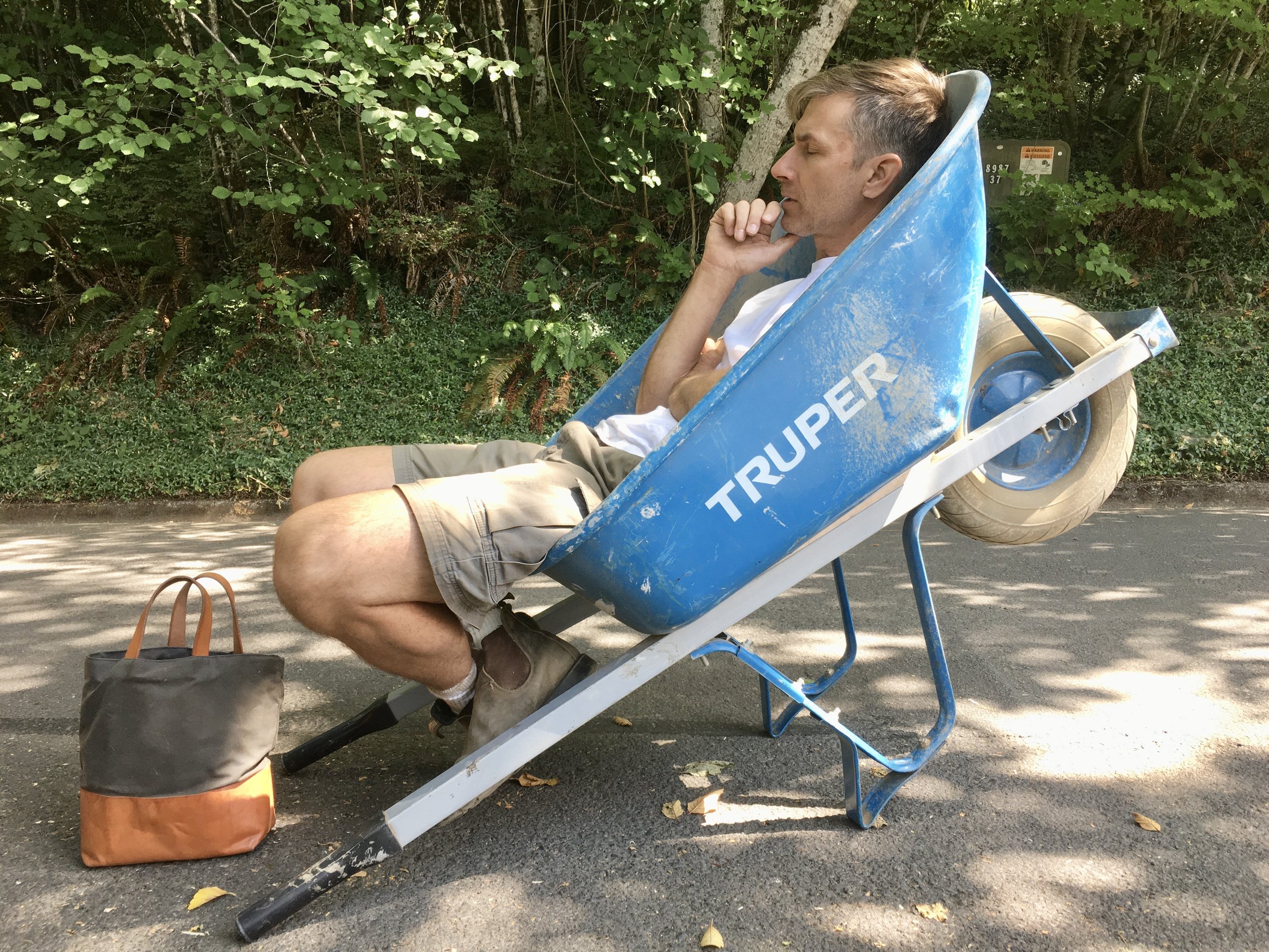 A man sitting in a blue wheelbarrow on a street, resting with his hand on his chin, surrounded by green trees and foliage.