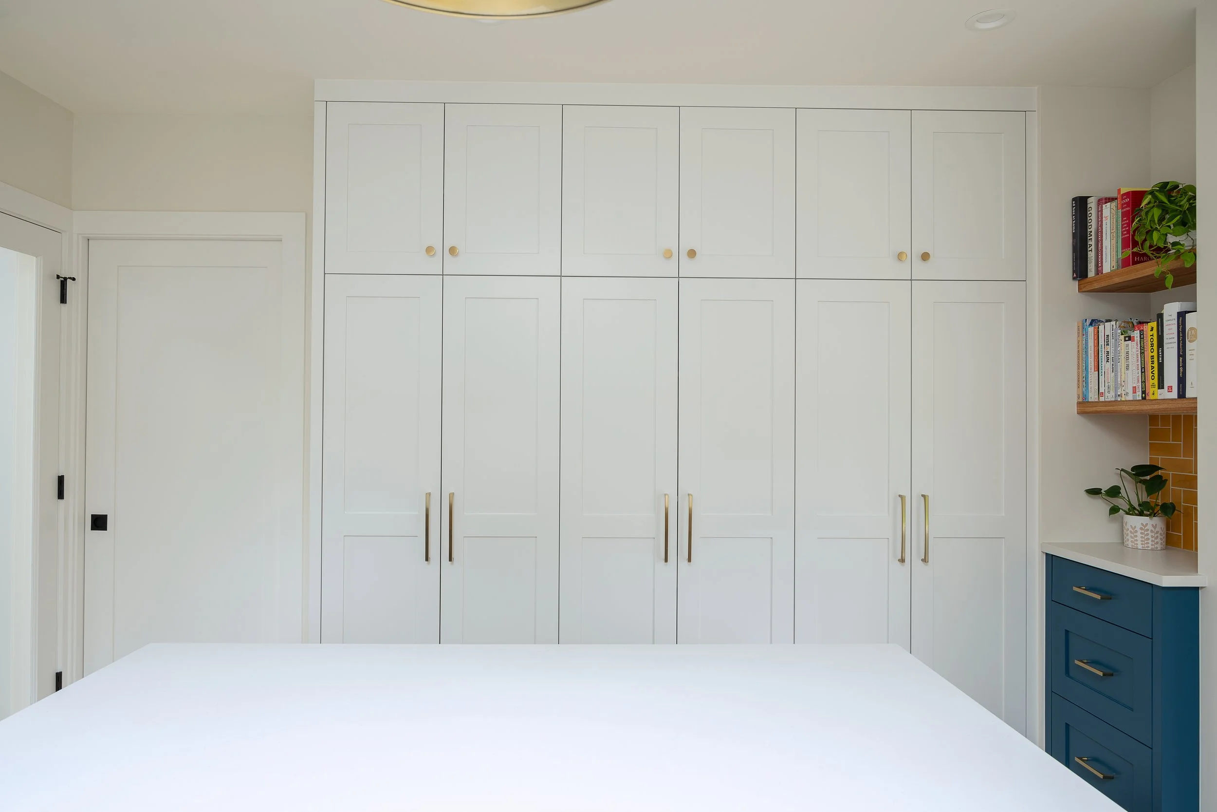 Minimalist remodeled white kitchen with built-in cabinets with gold knobs, a blue side cabinet with gold handles, a wooden shelf with books and a plant, and a white countertop.