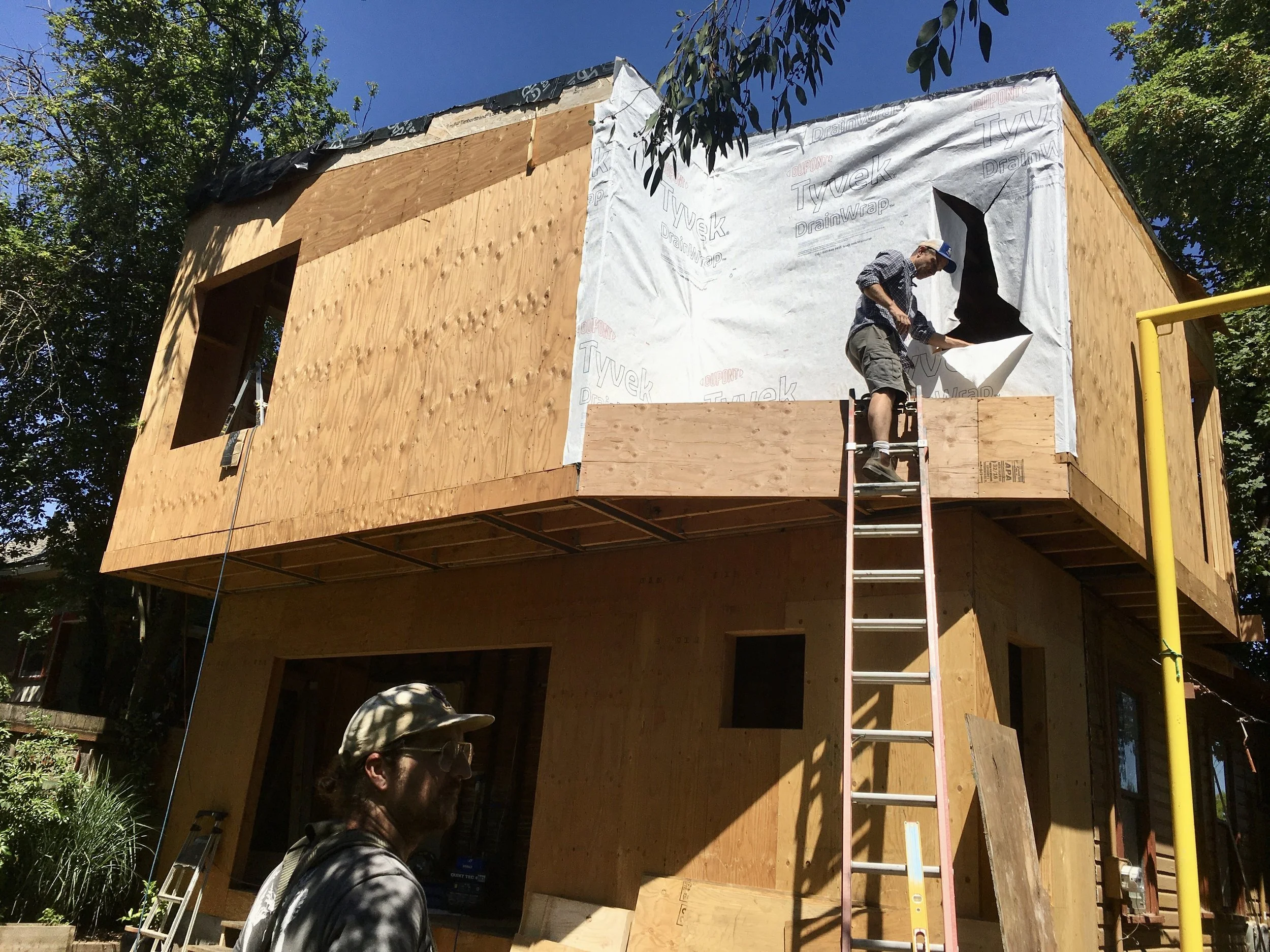 Construction site with a partially built wooden house, a man working on the side, ladder, and trees in the background, under a clear blue sky.